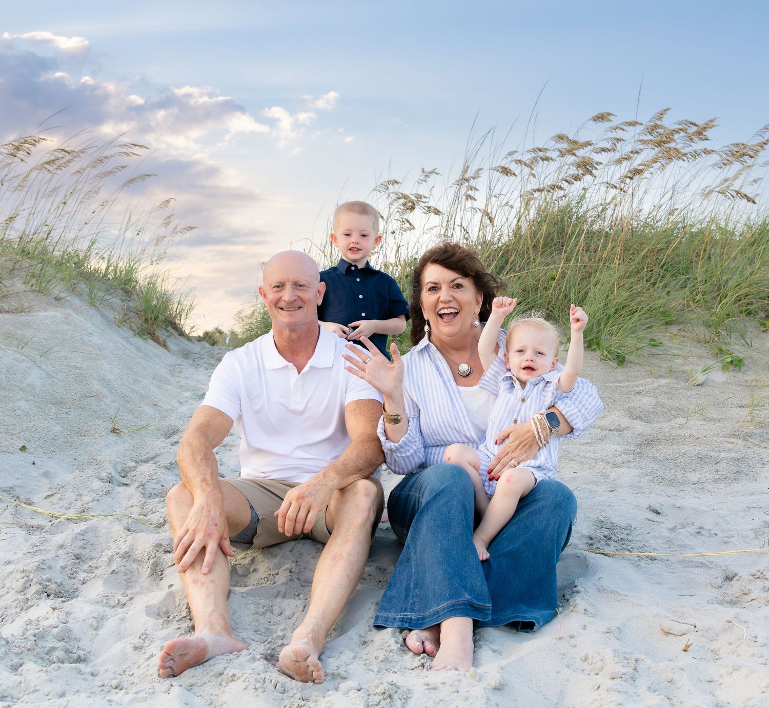 Happy grandparents sitting on sandy beach dunes with two young grandchildren, smiling outdoors at sunset.