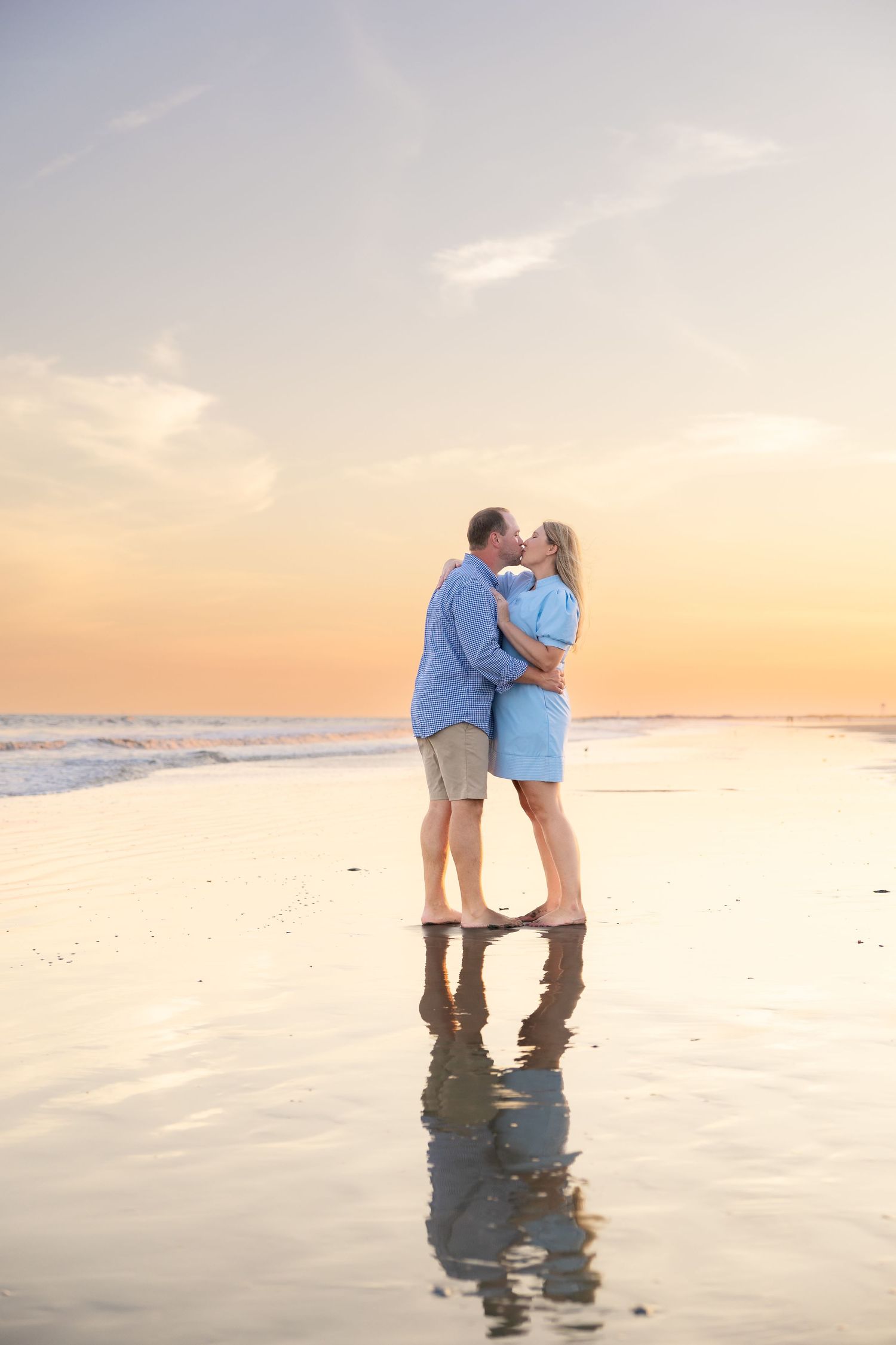 Couple sharing a romantic kiss on a wet beach at sunset, both wearing blue outfits with reflections in the sand.