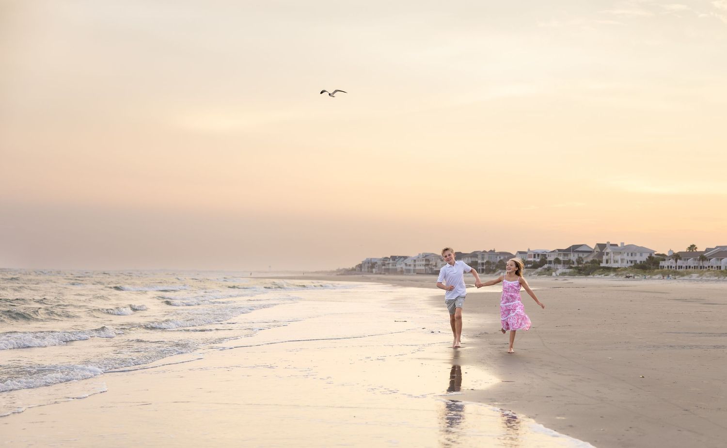 Couple holding hands running on a sandy beach at sunset with ocean waves and coastal town in background.