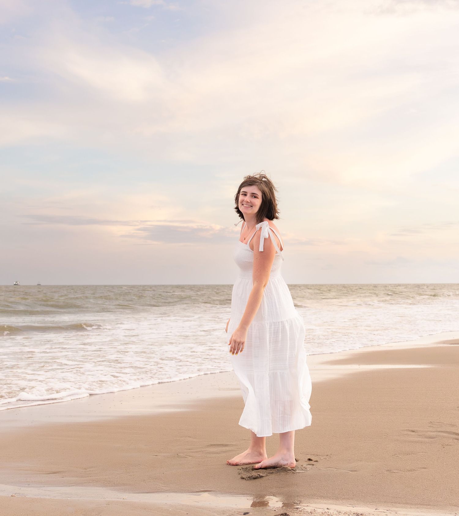 A young woman in a white dress stands barefoot on a sandy beach at sunset, smiling with ocean waves behind her.