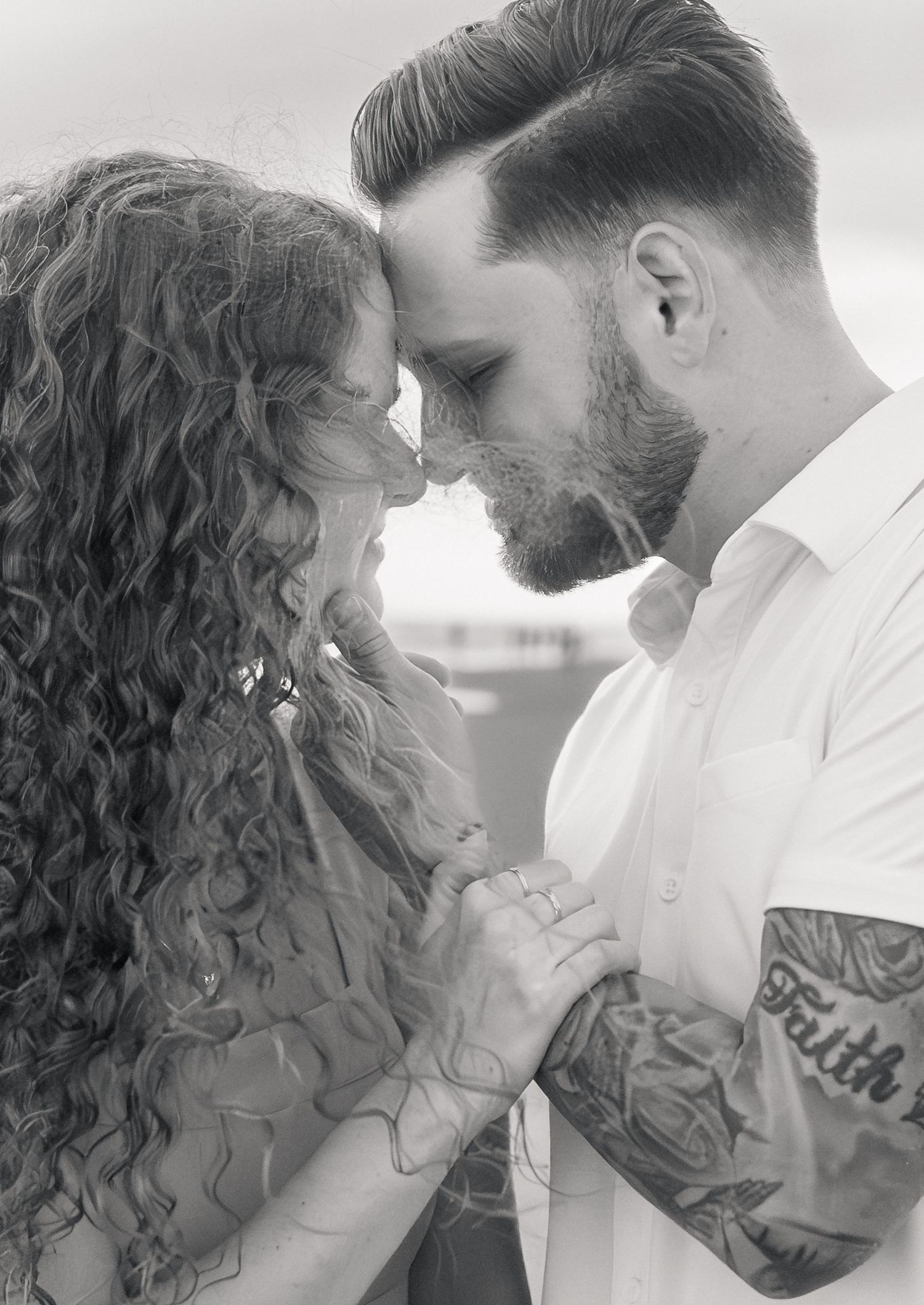 Black and white photo of a couple with foreheads touching, man with tattooed sleeve holding hands with curly-haired woman.