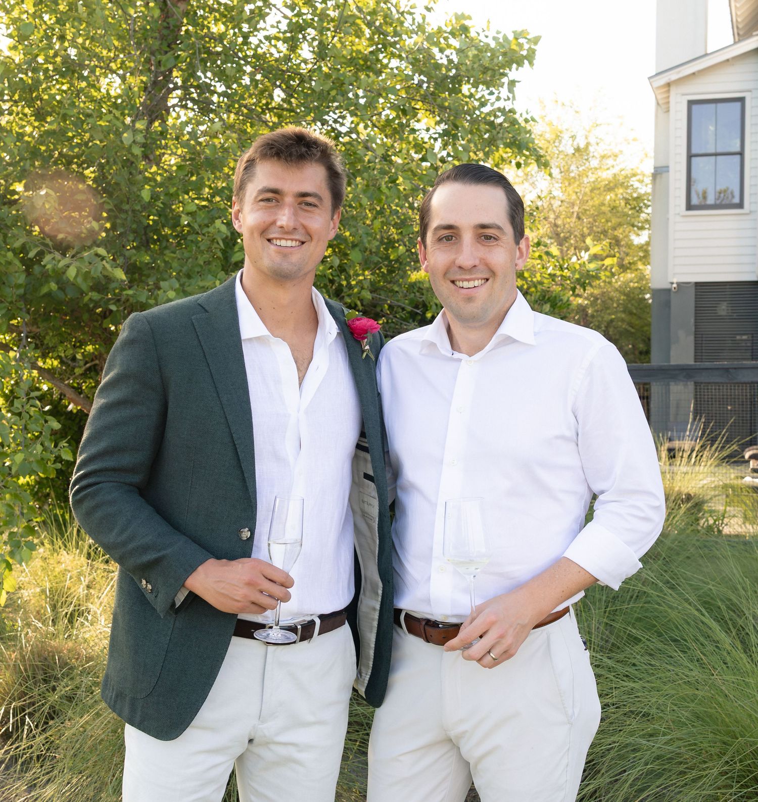 Two well-dressed men in blazers smile and hold wine glasses at an outdoor garden event.