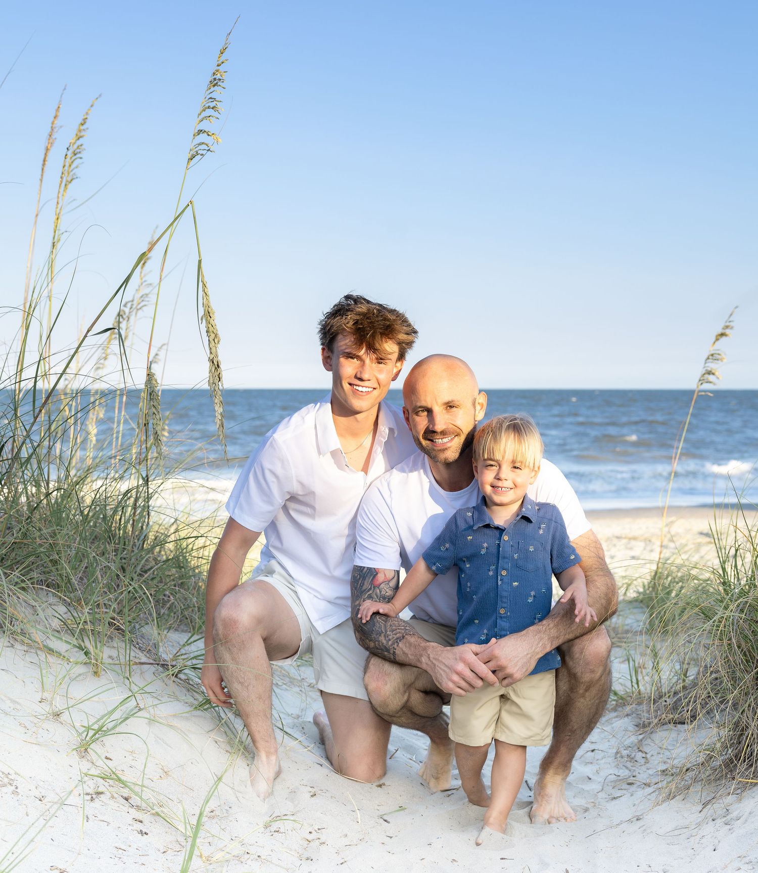 Two men and a young boy pose together on a sandy beach with sea oats and ocean in the background.