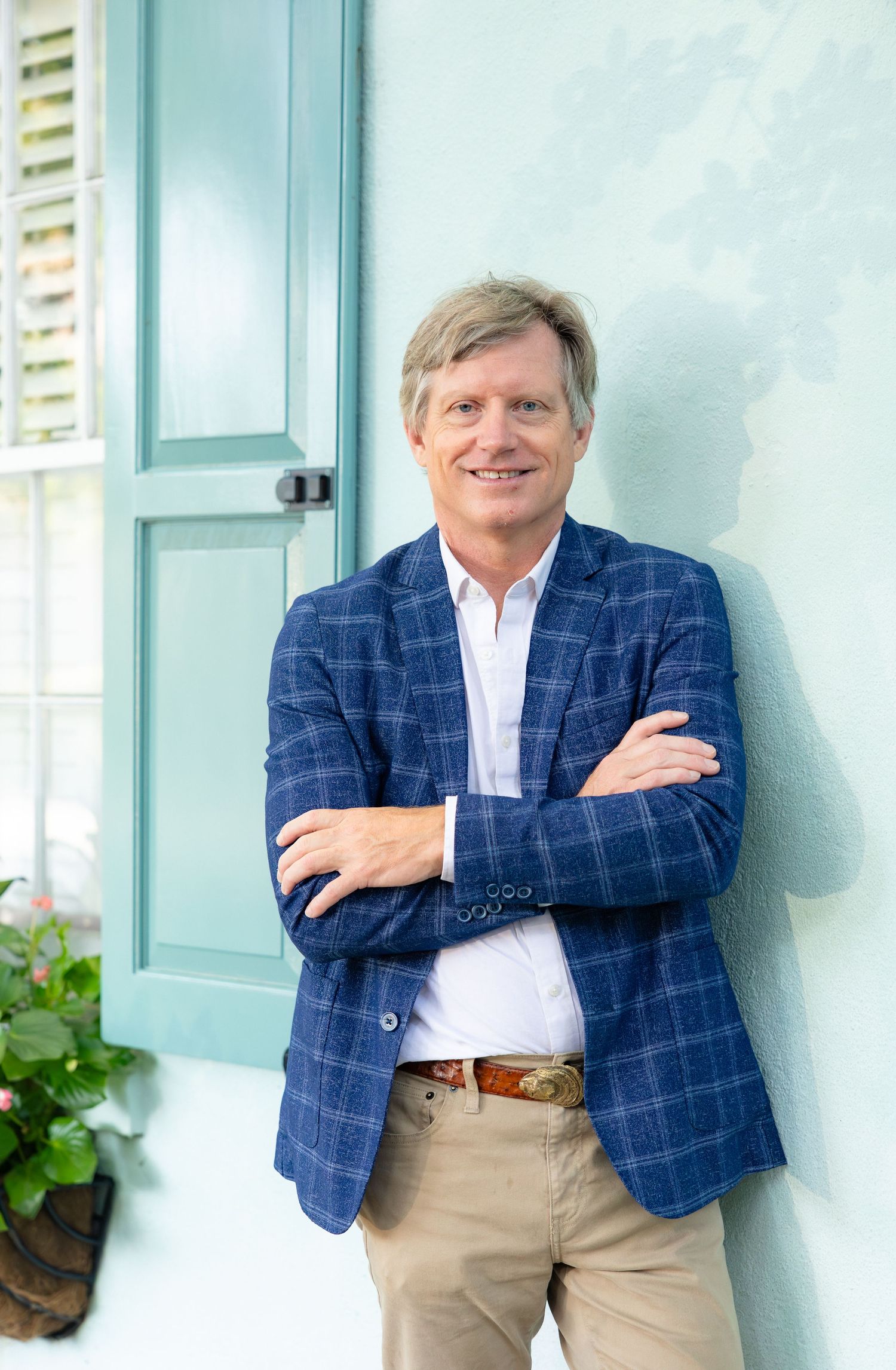 Man in blue plaid blazer smiling with arms crossed, leaning against a light blue wall near a teal door.