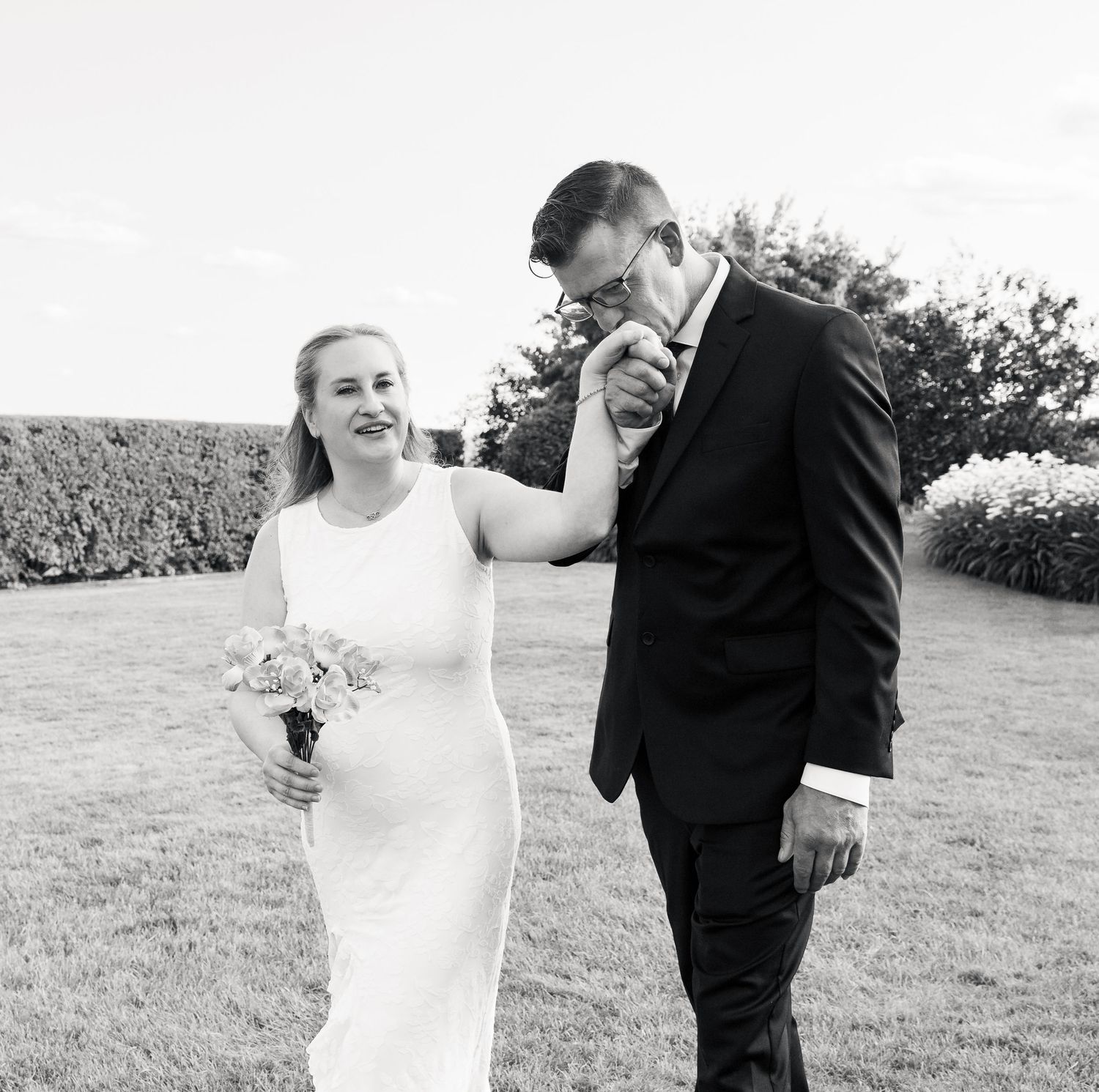Black and white photo of a groom kissing his bride's hand while she smiles holding a bouquet outdoors.