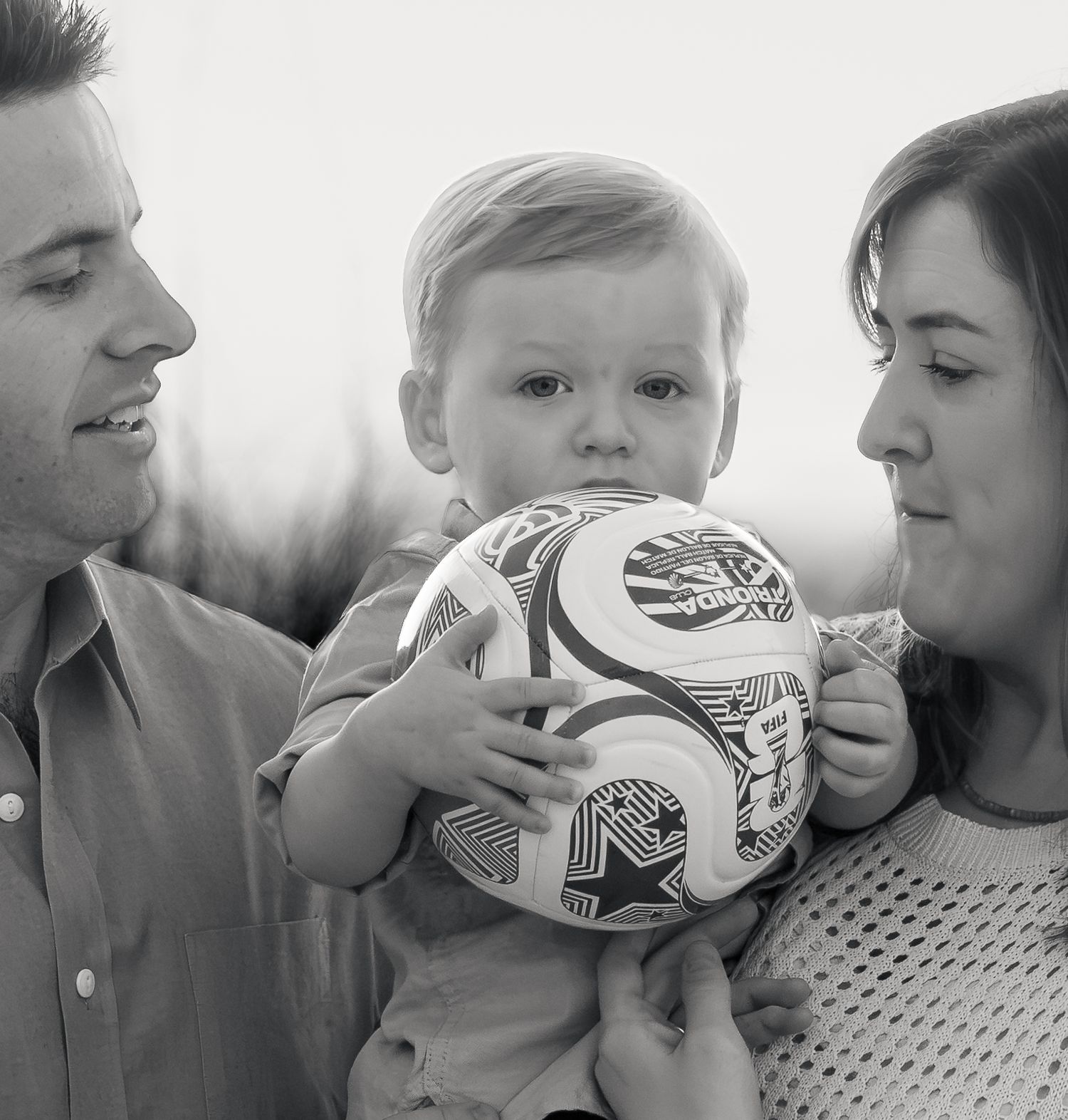 Black and white family portrait of a smiling man and woman holding a toddler who clutches a patterned soccer ball outdoors.