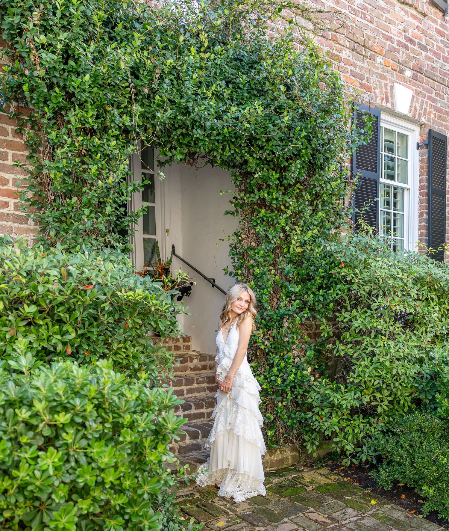 Woman in a white tiered ruffle dress stands before a brick doorway framed by lush green ivy and shrubs.