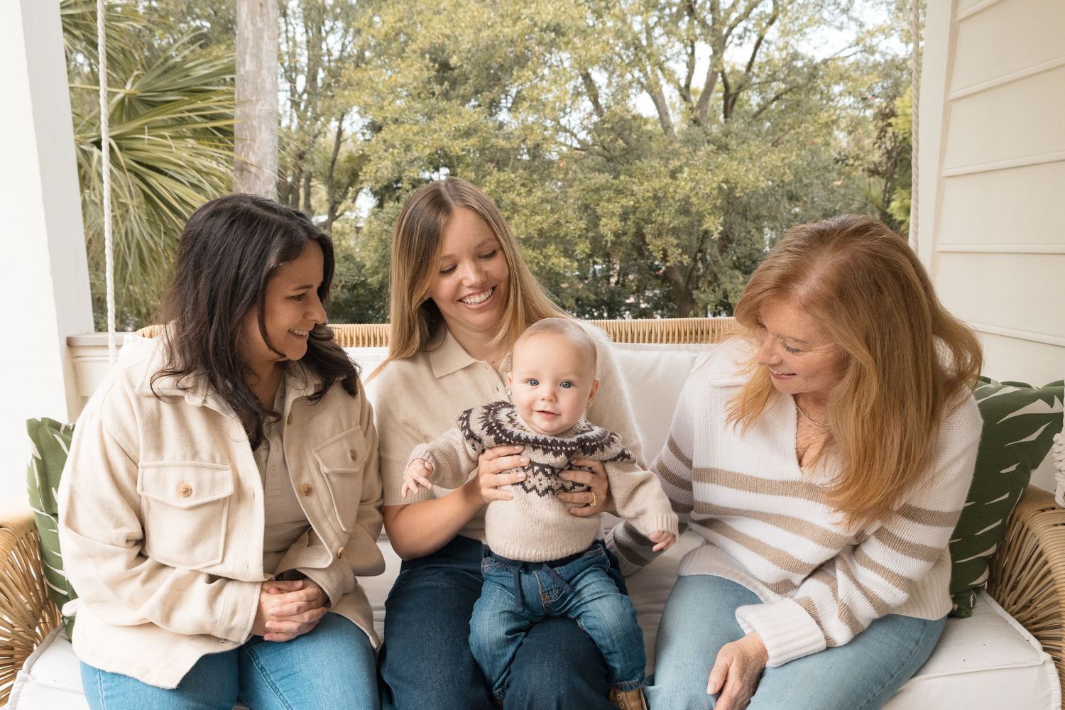 Three women sitting on a porch swing smiling at an adorable baby in a knit sweater surrounded by greenery.