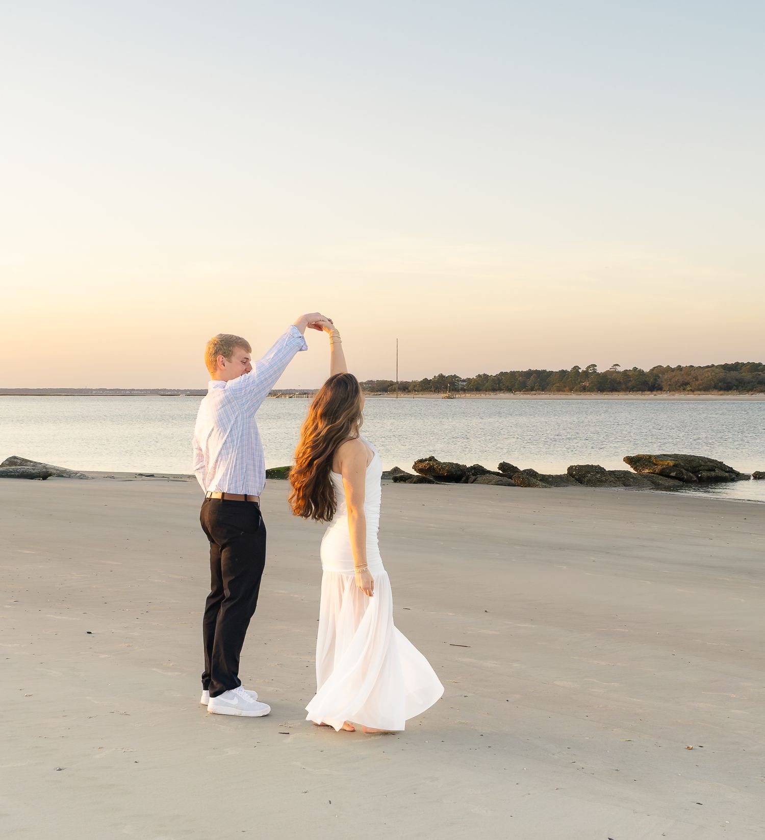 Couple dancing on a sandy beach at sunset, man in white shirt spinning woman in flowing white dress near the water.