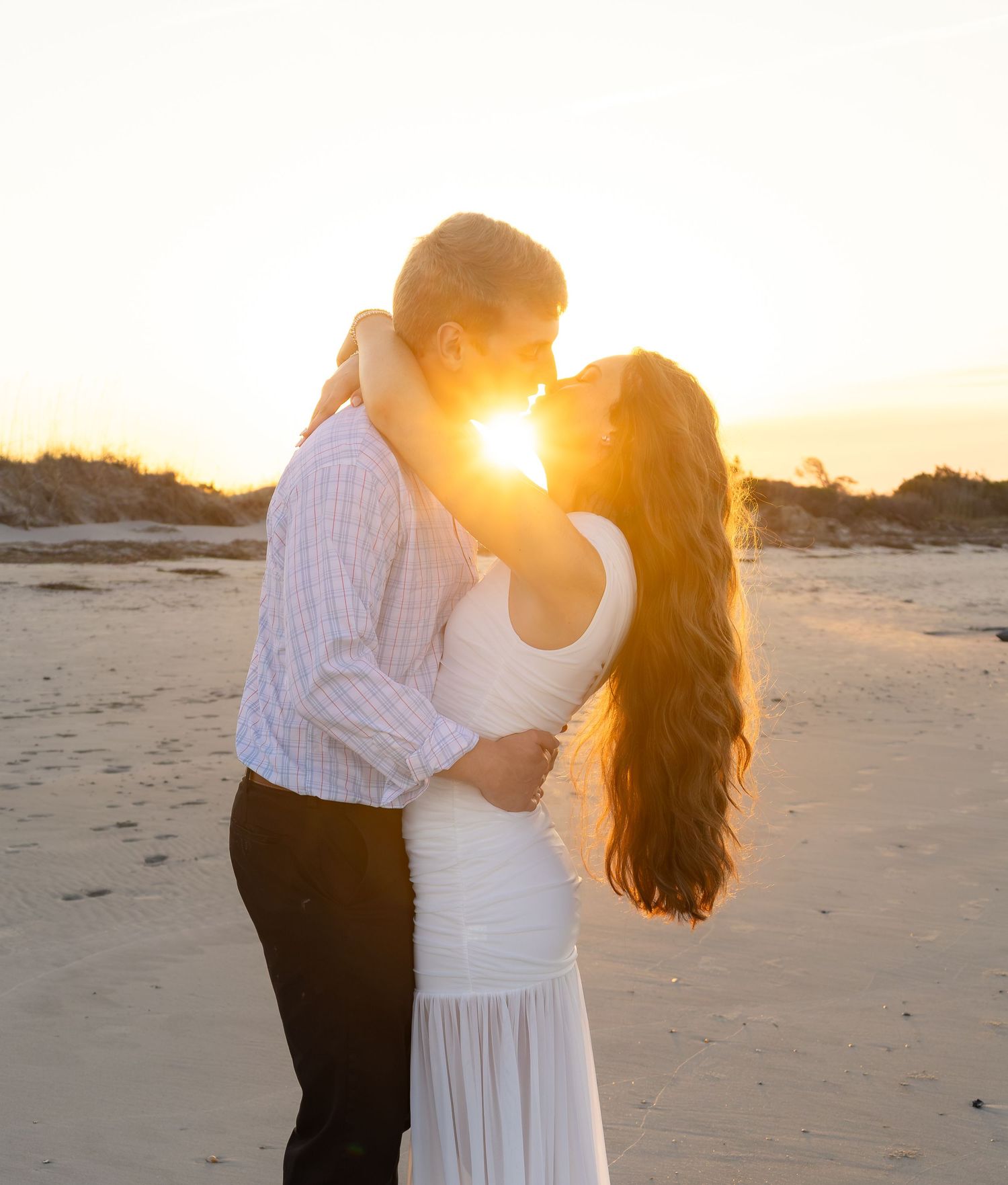 Couple sharing a romantic kiss on a sandy beach at golden sunset, woman in white dress with long dark hair.