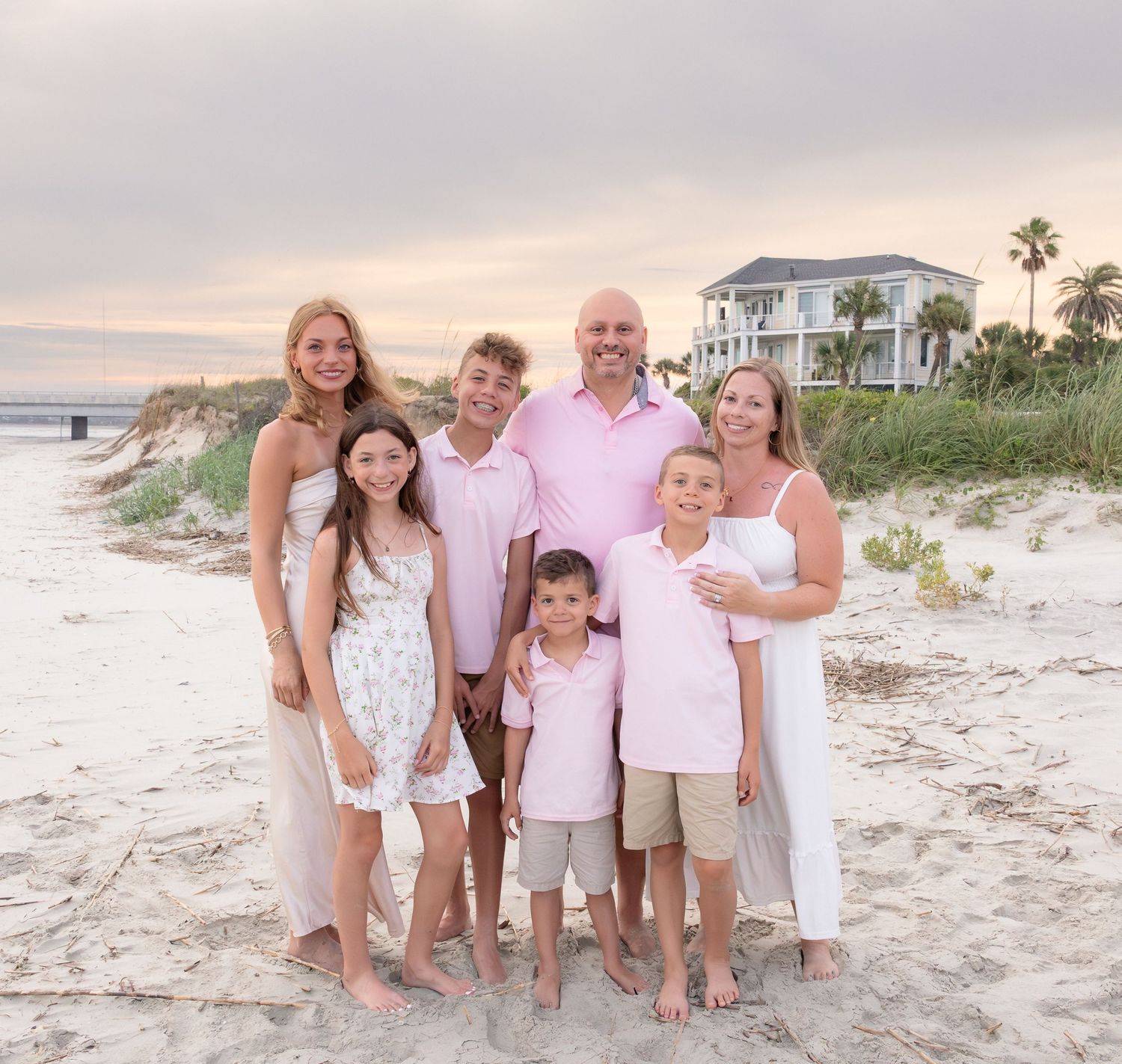 Family of seven posing together on a sandy beach at sunset with a coastal house and palm trees in background.