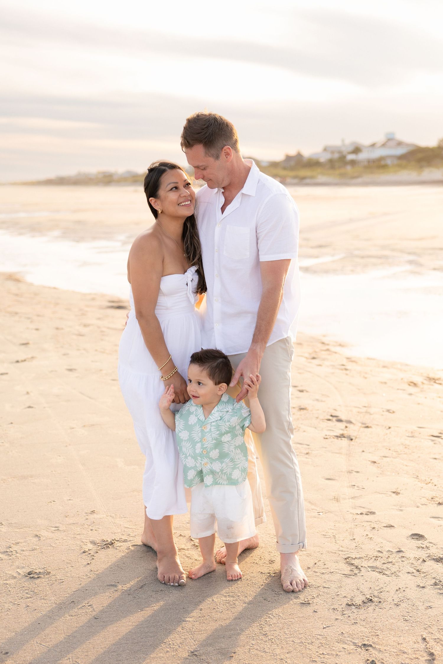 Family of three posing on a sandy beach at sunset, parents in white outfits holding hands with their toddler son.
