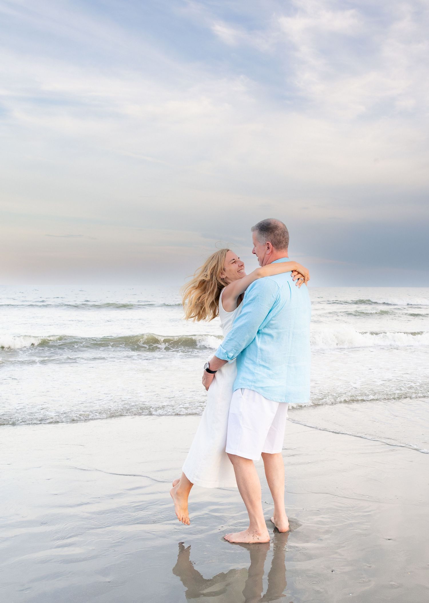 Couple embracing on a sandy beach at sunset, woman lifted in man's arms, ocean waves in the background.