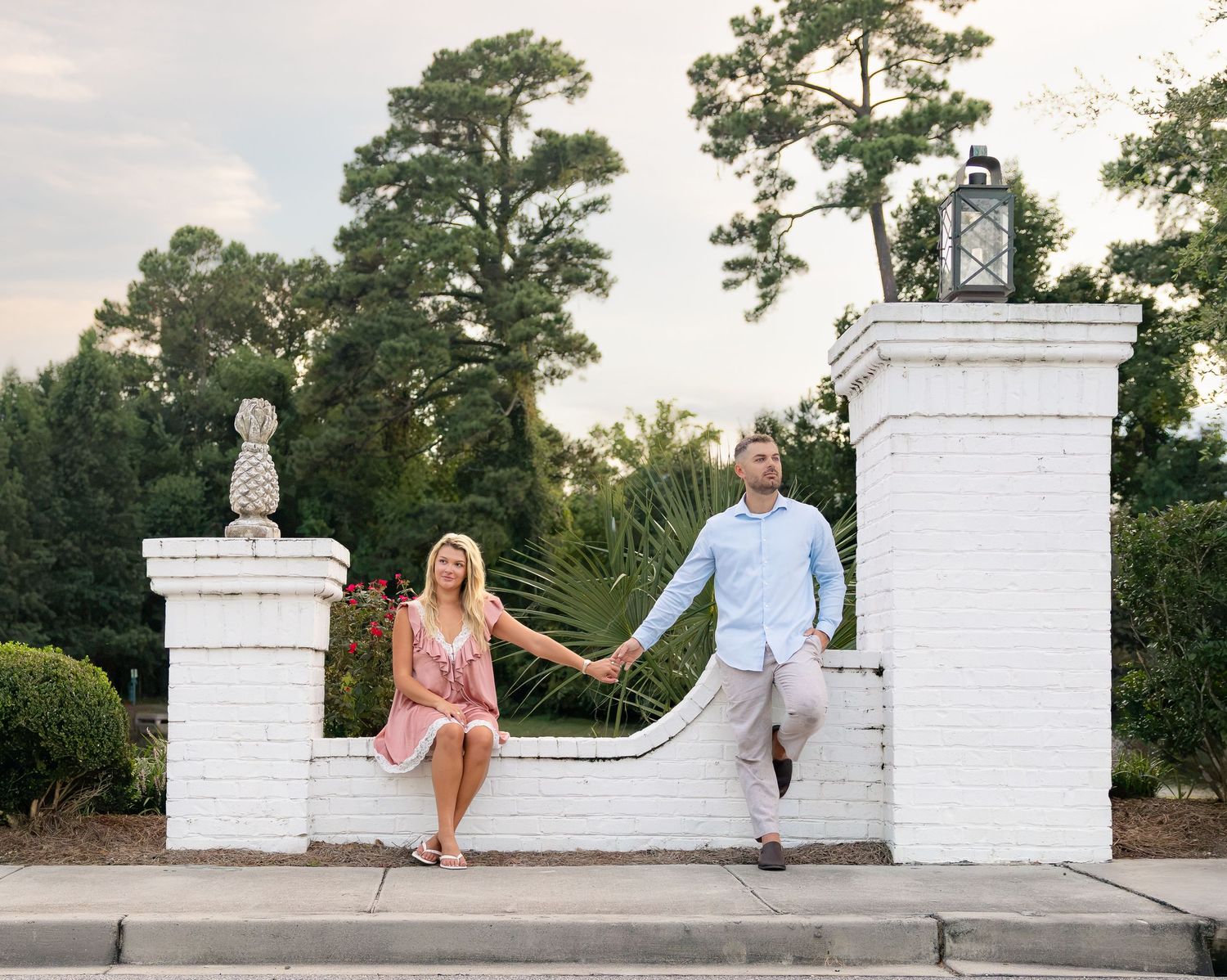Couple holding hands on a white brick garden wall with pineapple finials and lantern, surrounded by lush greenery.