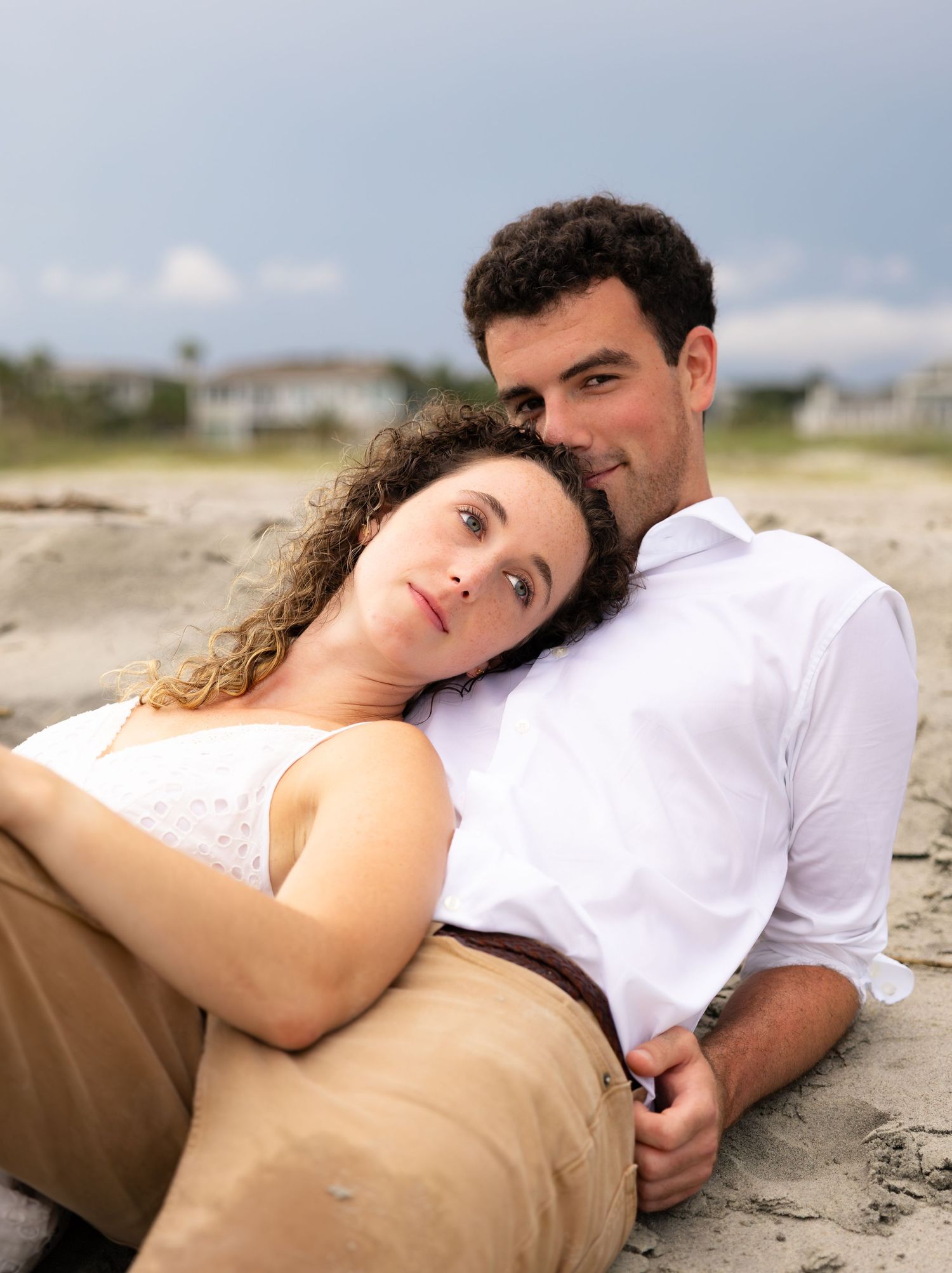 Romantic couple sitting together on a sandy beach, woman resting head on man's shoulder, cloudy sky backdrop.
