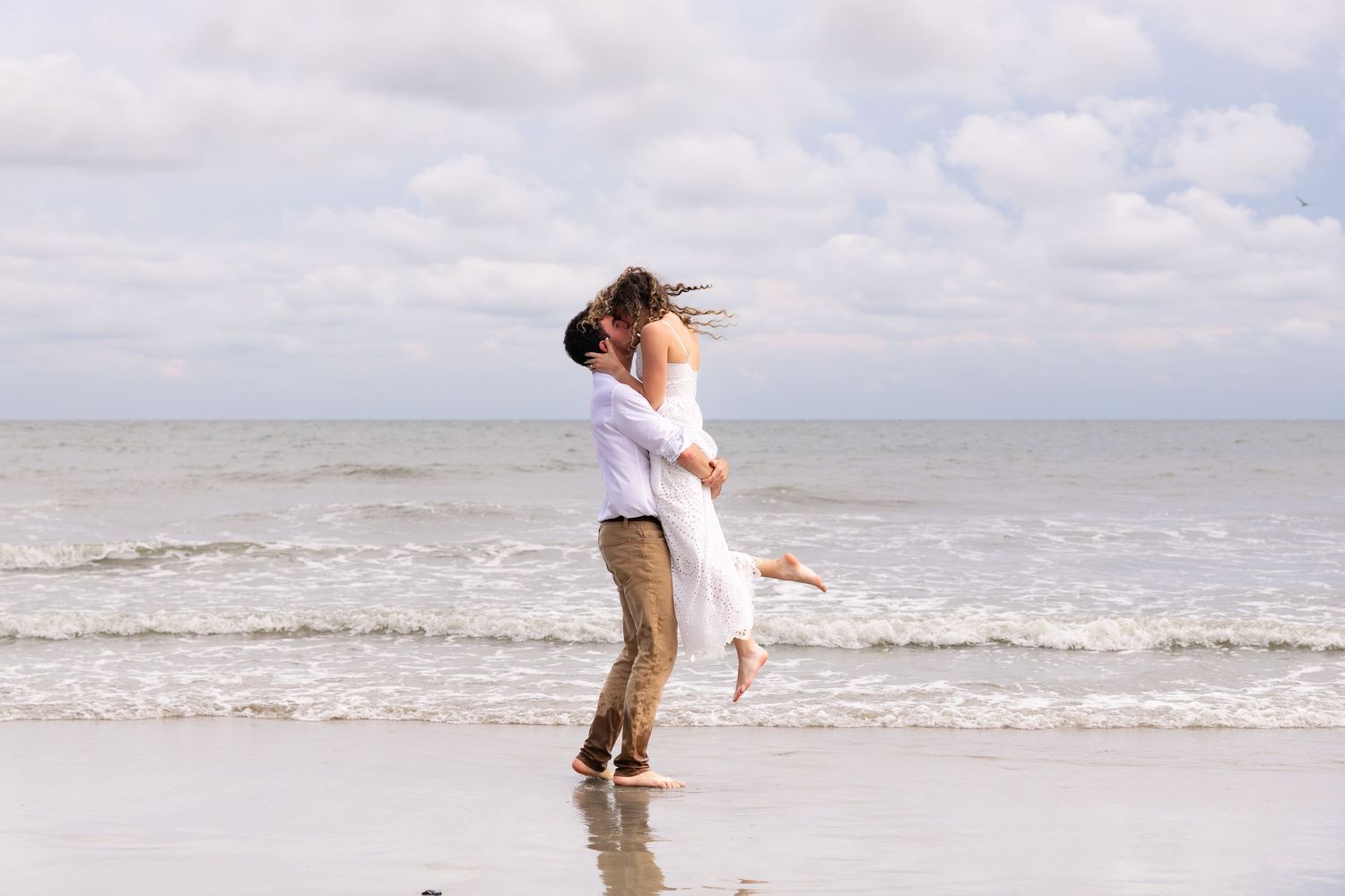 Romantic couple embracing on a beach, man lifting woman in white dress near ocean waves under cloudy sky.