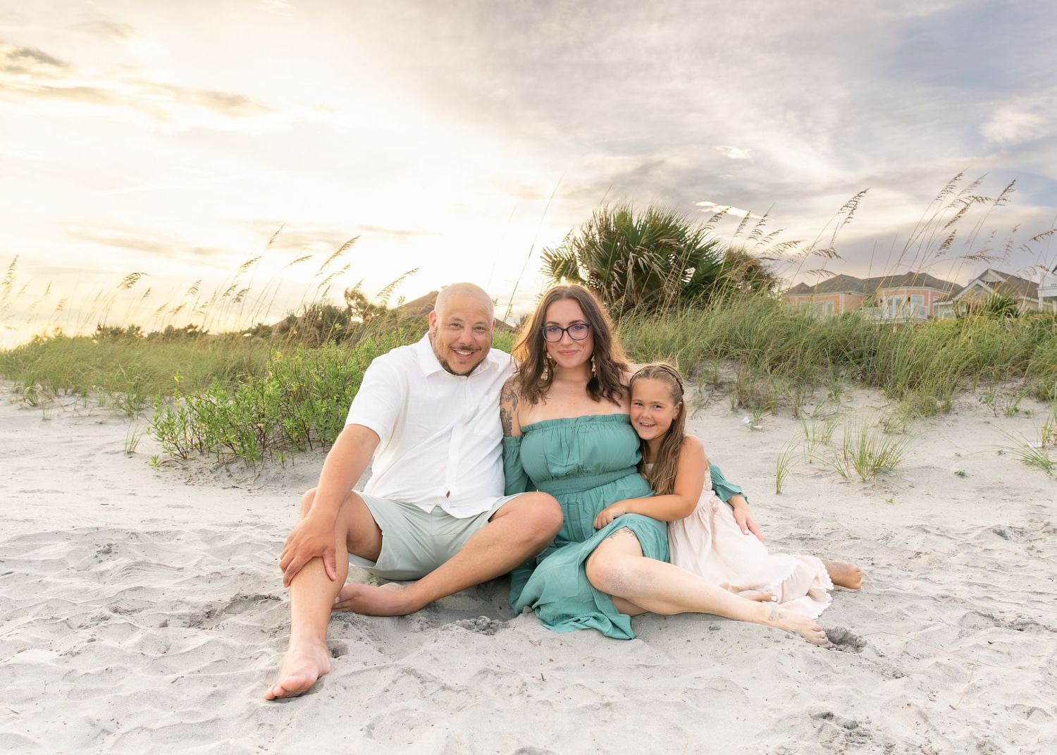 Family of three sitting on beach sand at golden hour with sea oats and coastal homes behind them.