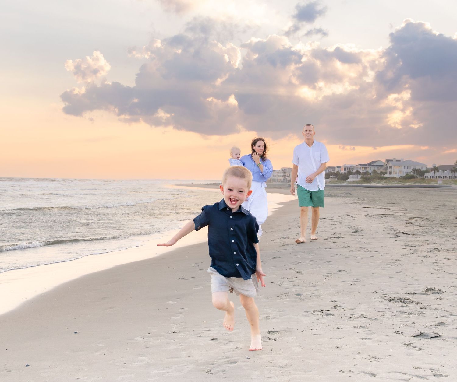 A young boy runs on a beach at sunset while his family watches in the background near the ocean.