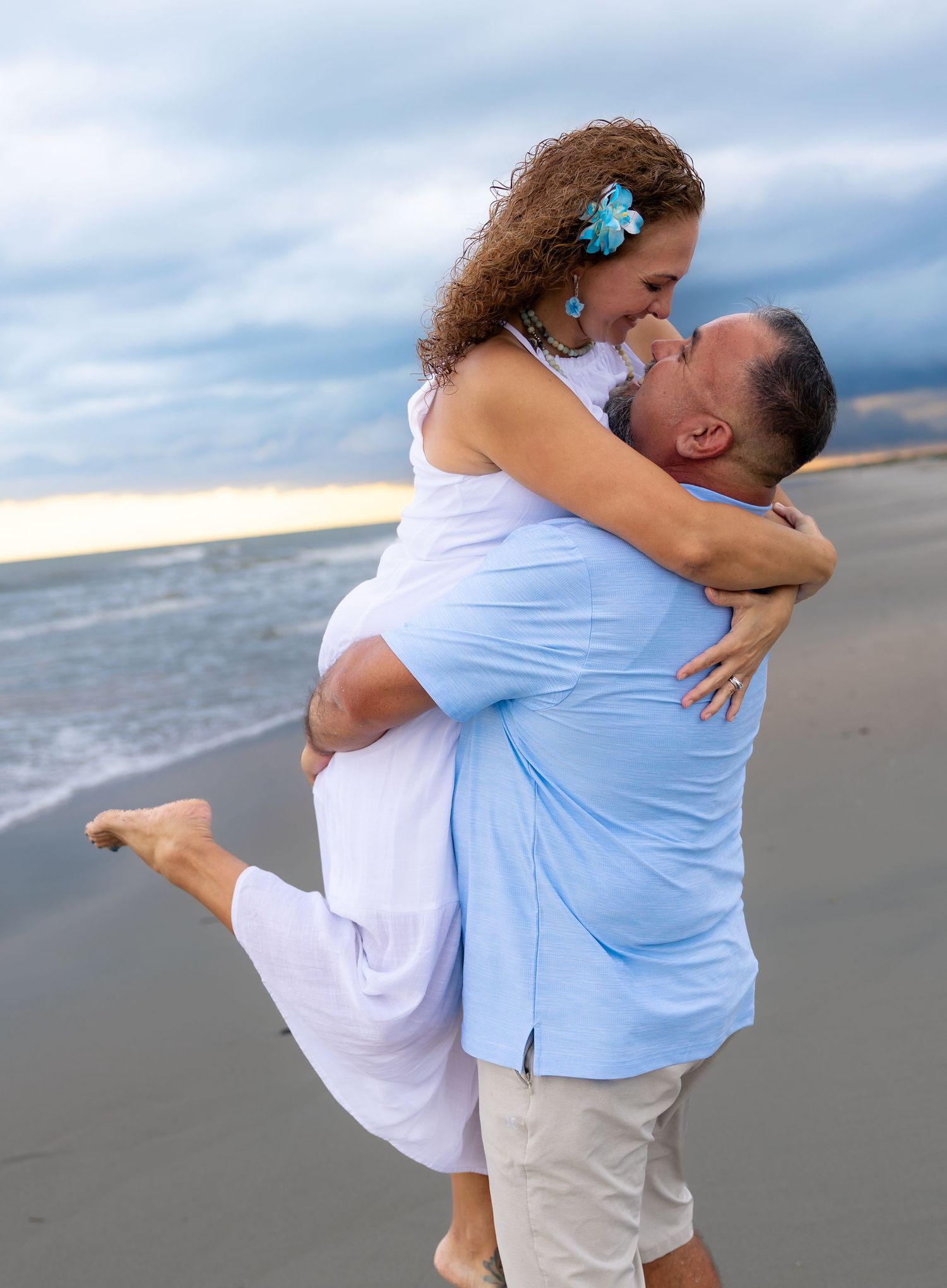 Couple sharing a romantic kiss on the beach, man lifting woman in white dress with turquoise accessories.