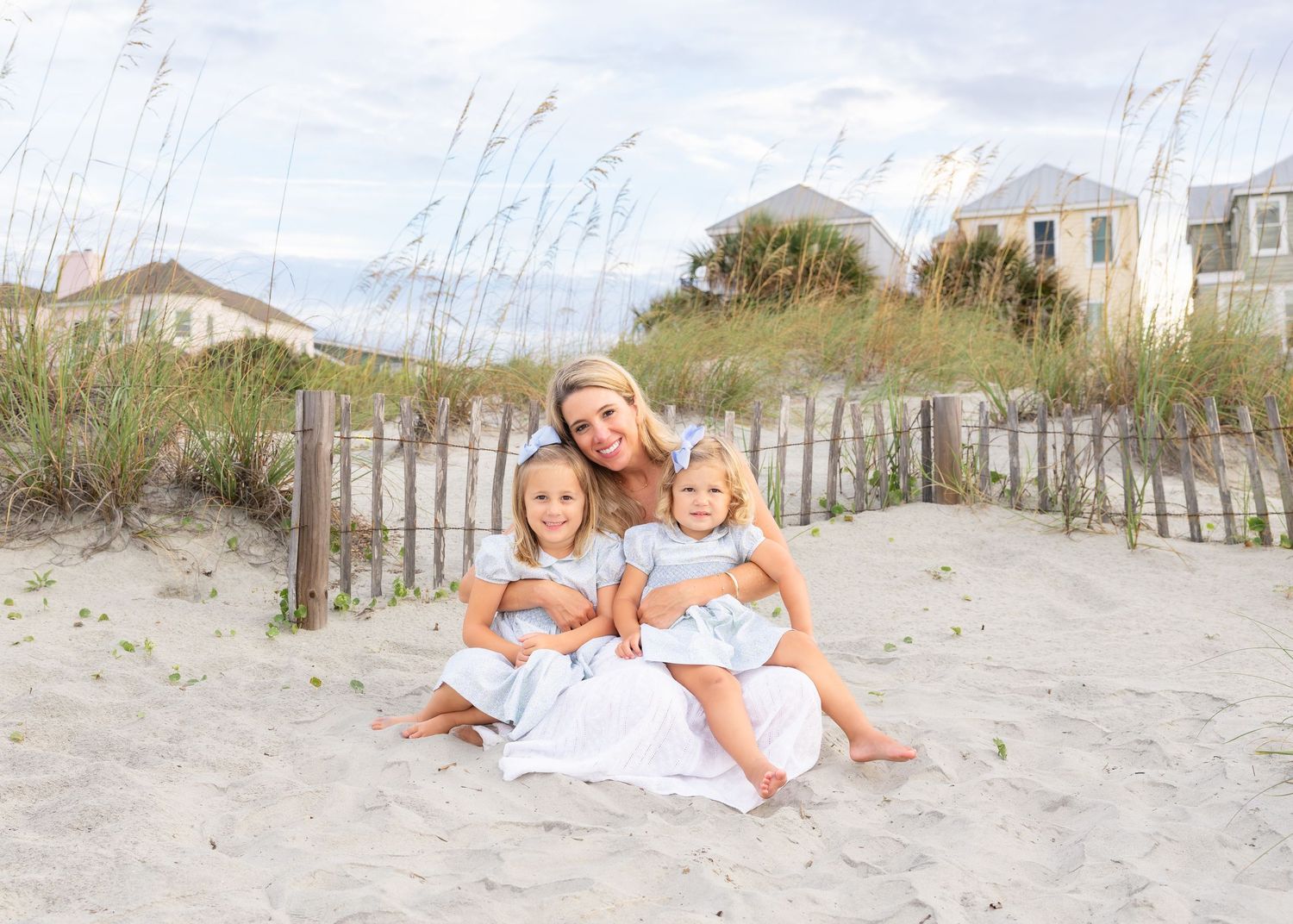 A mother and two young daughters in matching white dresses smile together on a sandy beach near dunes.
