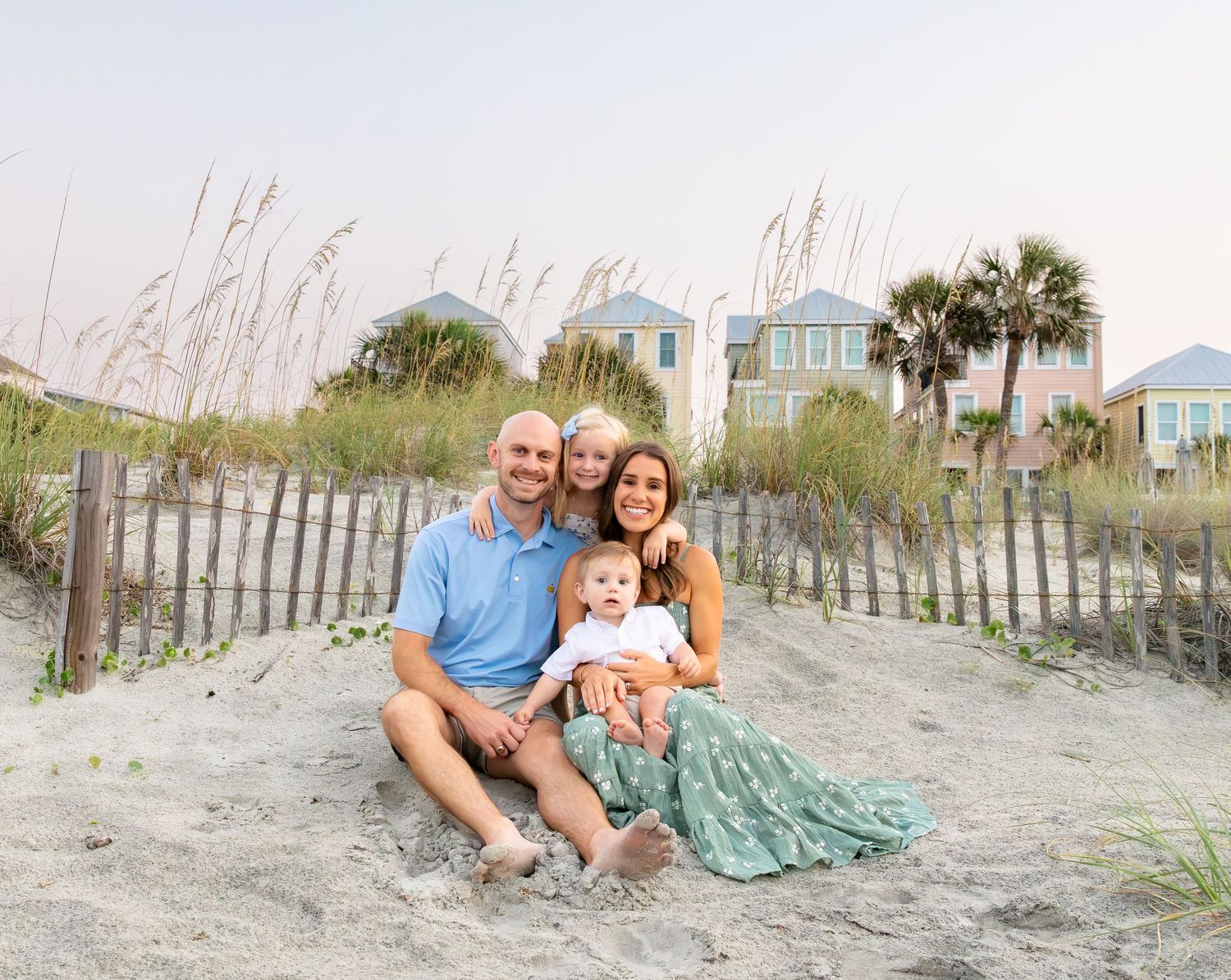 Family of four posing on sandy beach dunes with colorful beach houses and palm trees in background.
