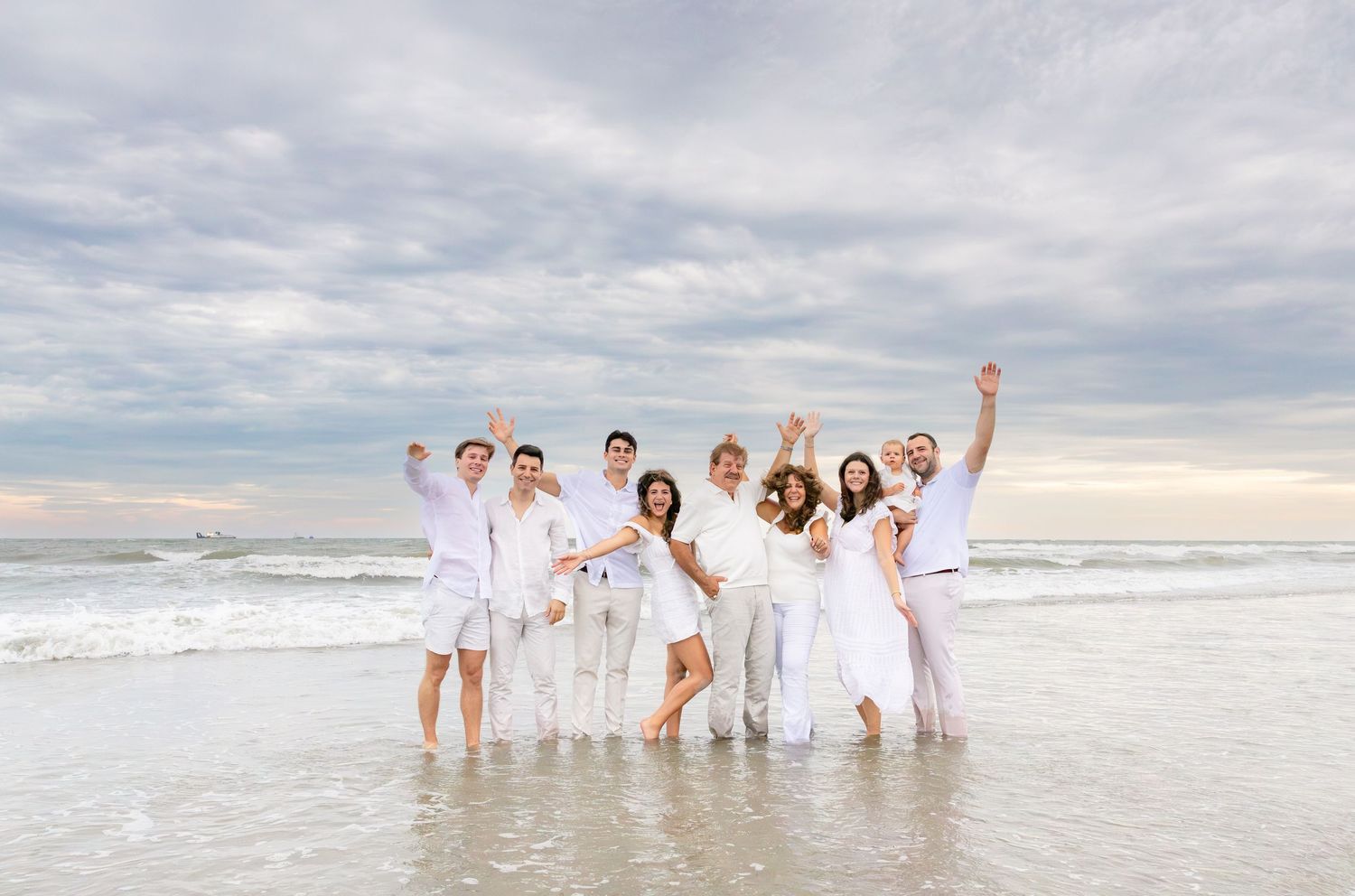 Group of eight people dressed in white celebrating on a sandy beach with ocean waves in the background.
