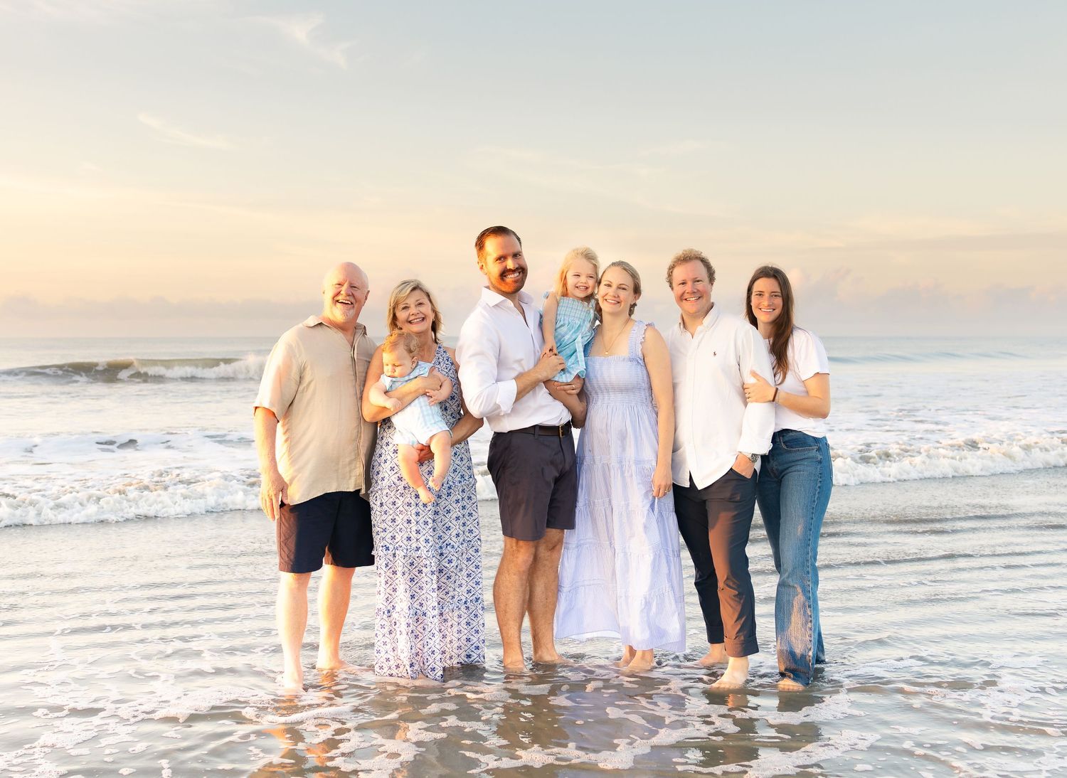 Large family group smiling together on a sunny beach with ocean waves, capturing a joyful multigenerational portrait.