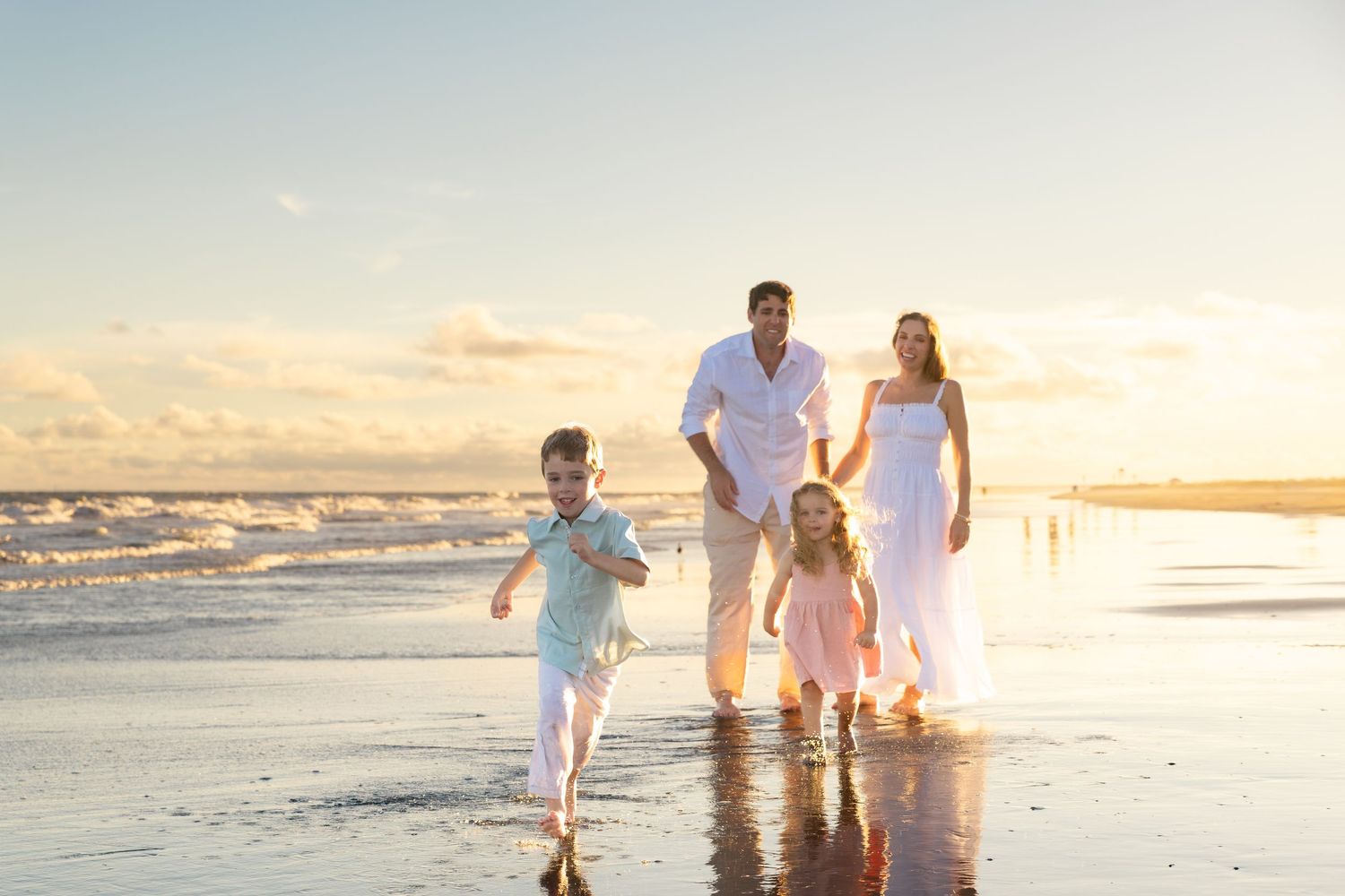 Family of four enjoying a sunset beach walk, with children running along the shoreline in white summer outfits.