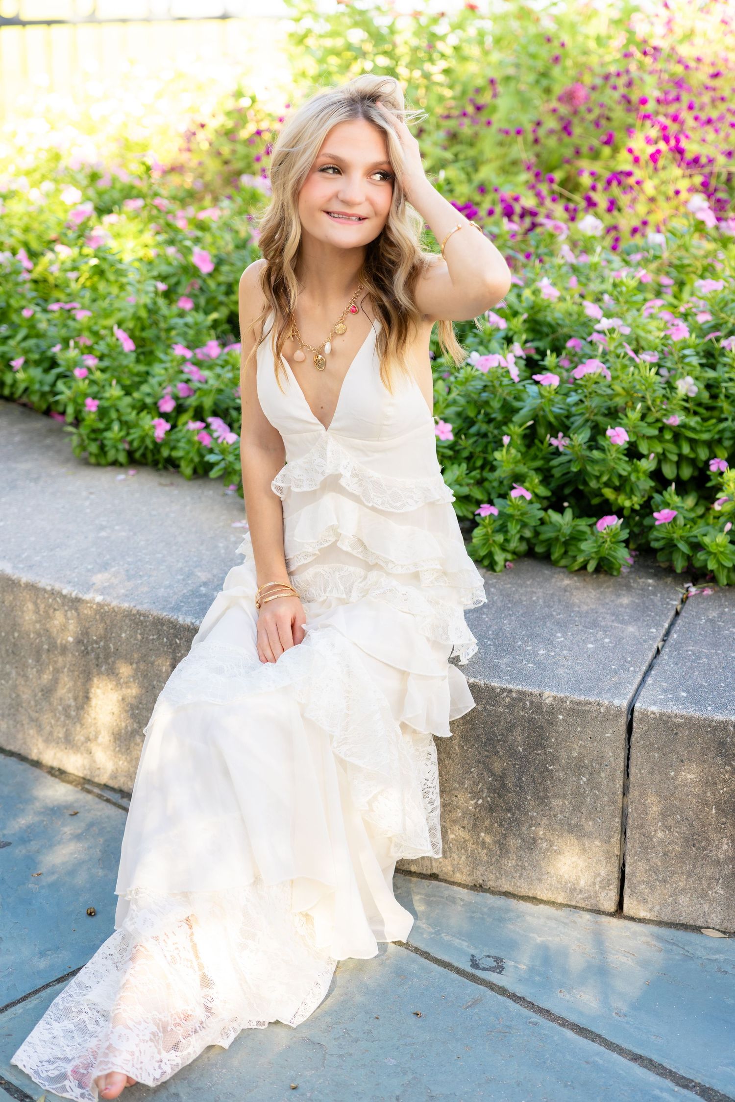 Young woman in white ruffled lace maxi dress sitting on stone steps surrounded by colorful garden flowers.