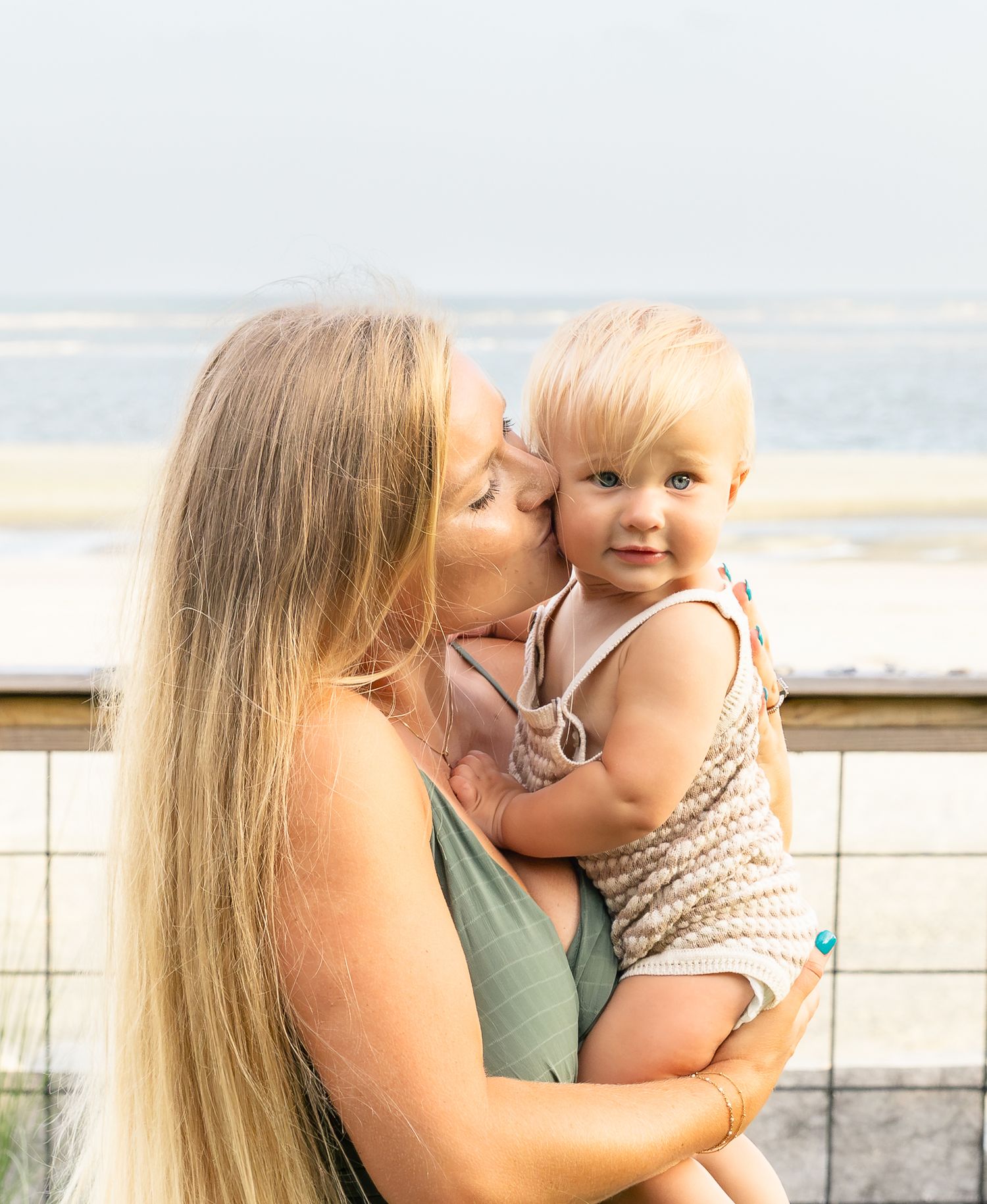 A woman with long blonde hair kisses a smiling toddler on the cheek while holding the child on a beachside boardwalk overlooking the ocean.