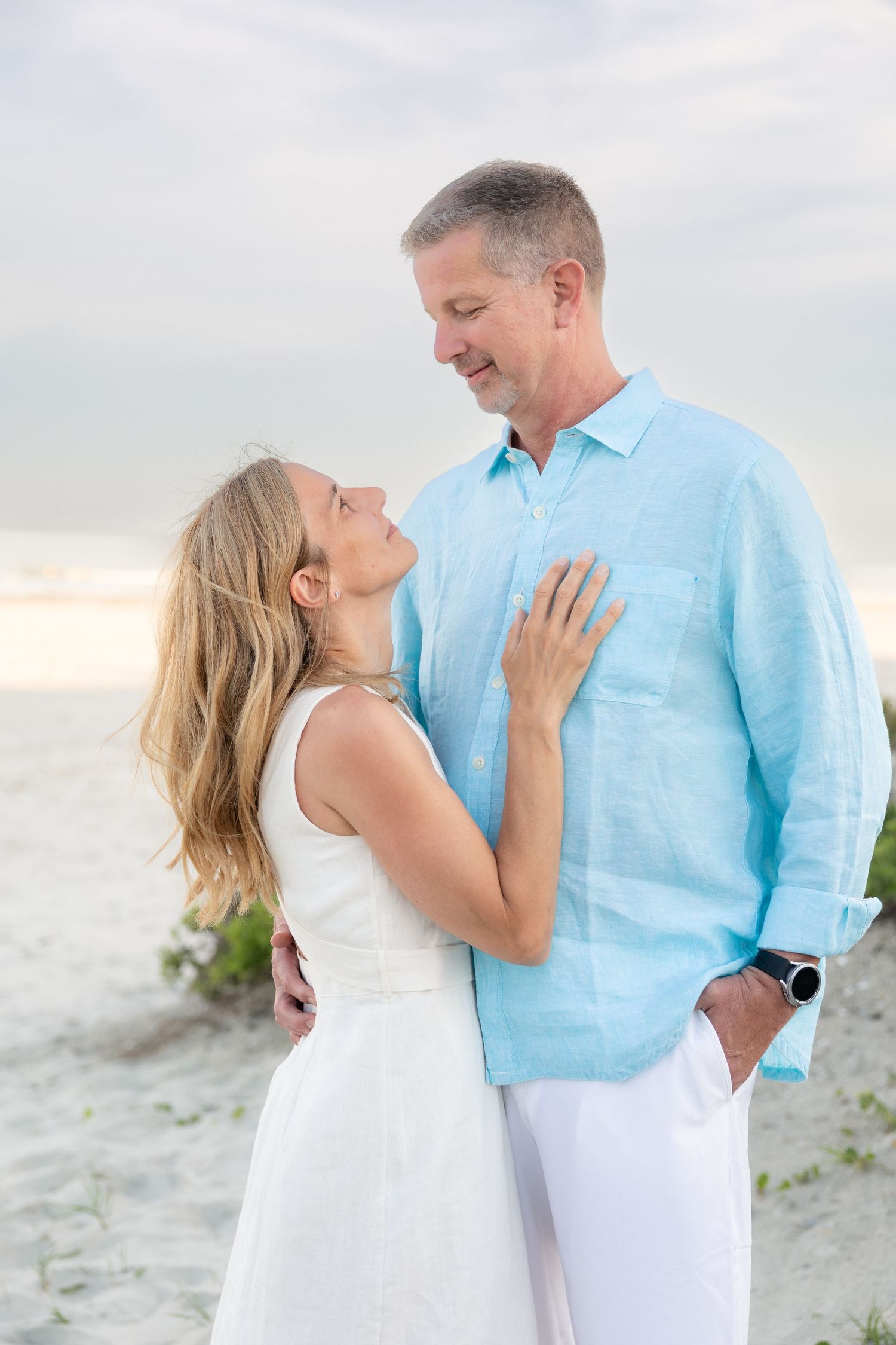 Couple embracing on a sandy beach, woman in white dress, man in light blue shirt, sharing a romantic moment.