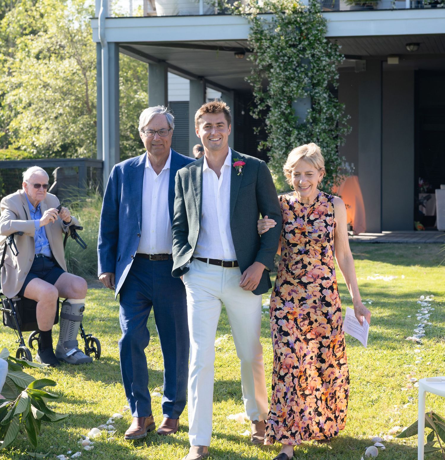 Groom walks with parents across green lawn at outdoor wedding ceremony near modern building.