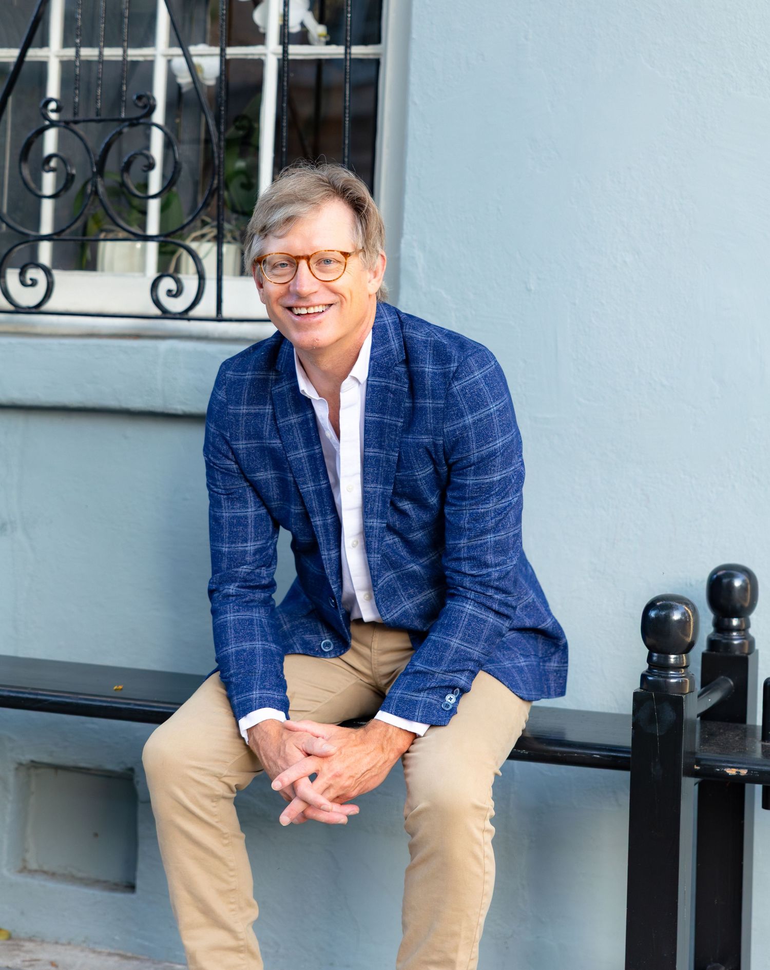Smiling middle-aged man in blue plaid blazer and khakis sitting outdoors on a ledge near a light blue building.