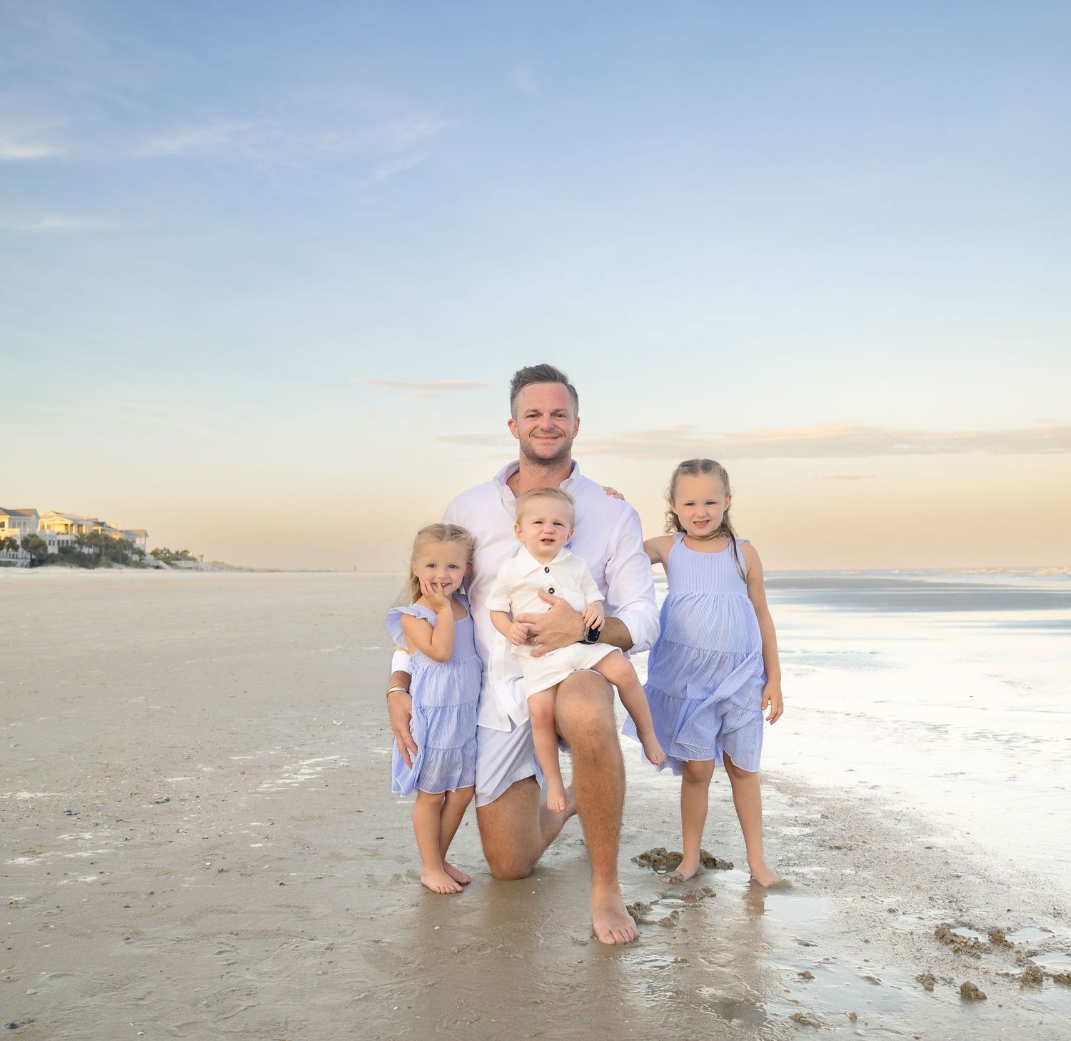 A father kneels on a sandy beach with three young children at sunset, smiling for a family portrait.