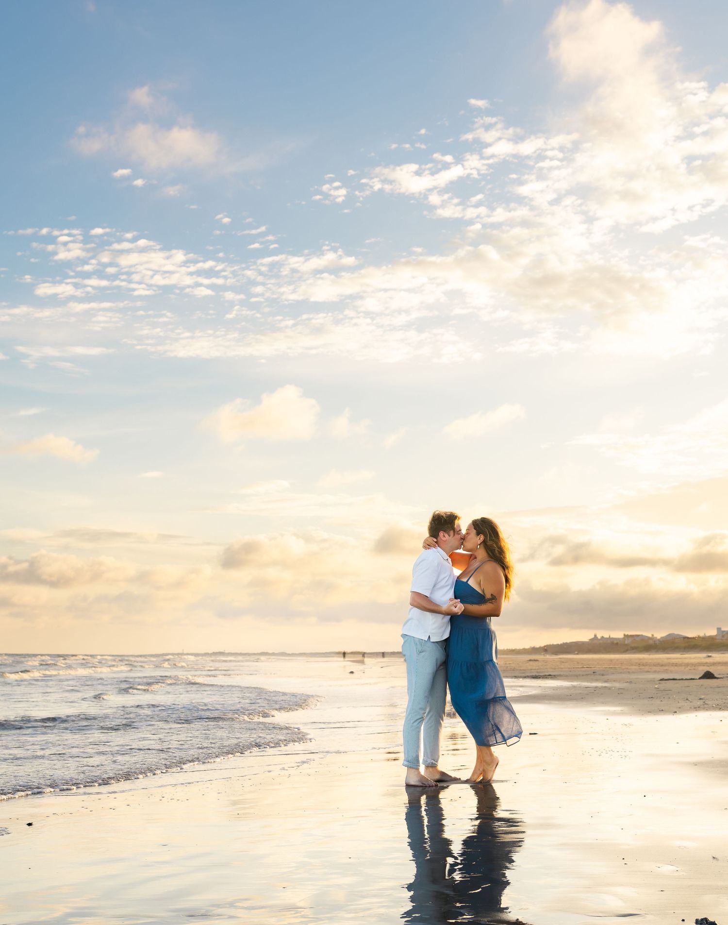 Couple sharing a romantic kiss on a reflective beach at sunset, with ocean waves and a colorful cloudy sky.