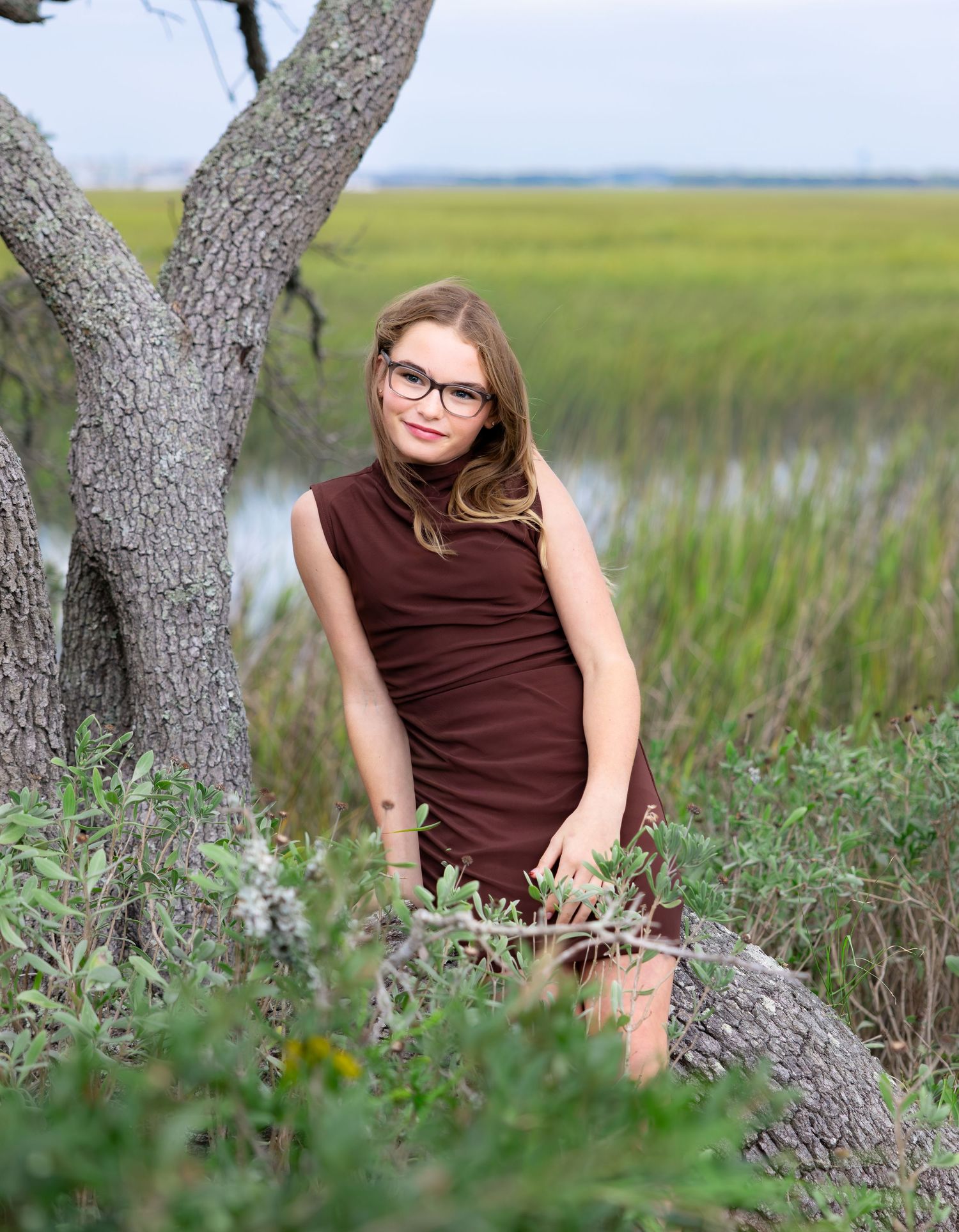 Young woman with glasses and long hair, wearing a brown sleeveless dress, sits beside a gnarled tree overlooking a green marsh.