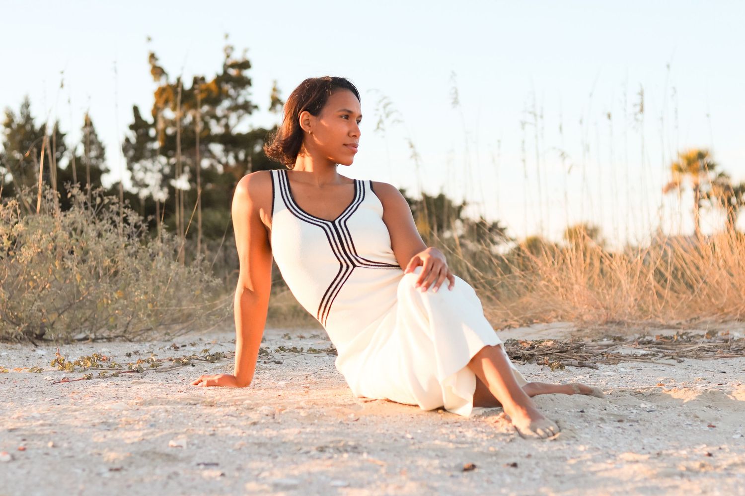Woman in white dress sitting gracefully on sandy beach with golden grasses and palm trees in background.