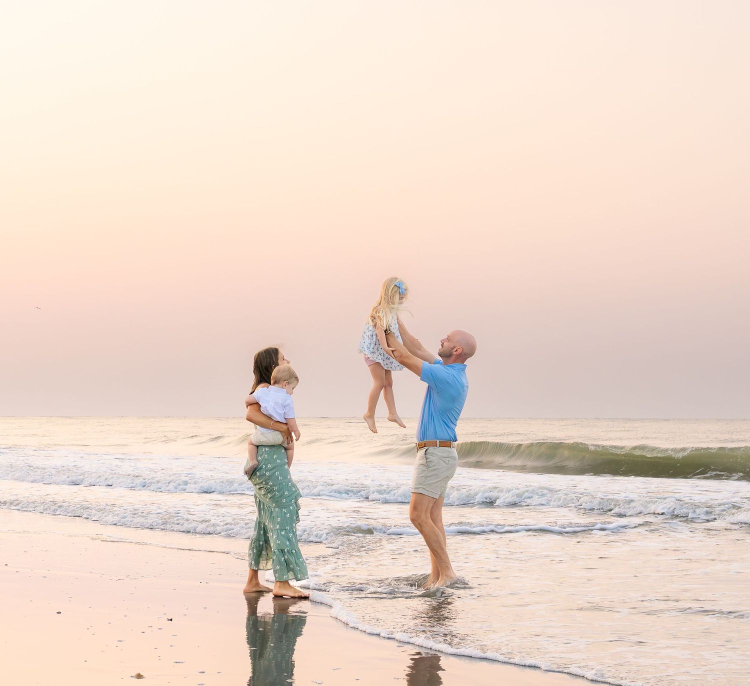 Family enjoying a beach sunset, parents playing with two young children at the water's edge on a serene shoreline.