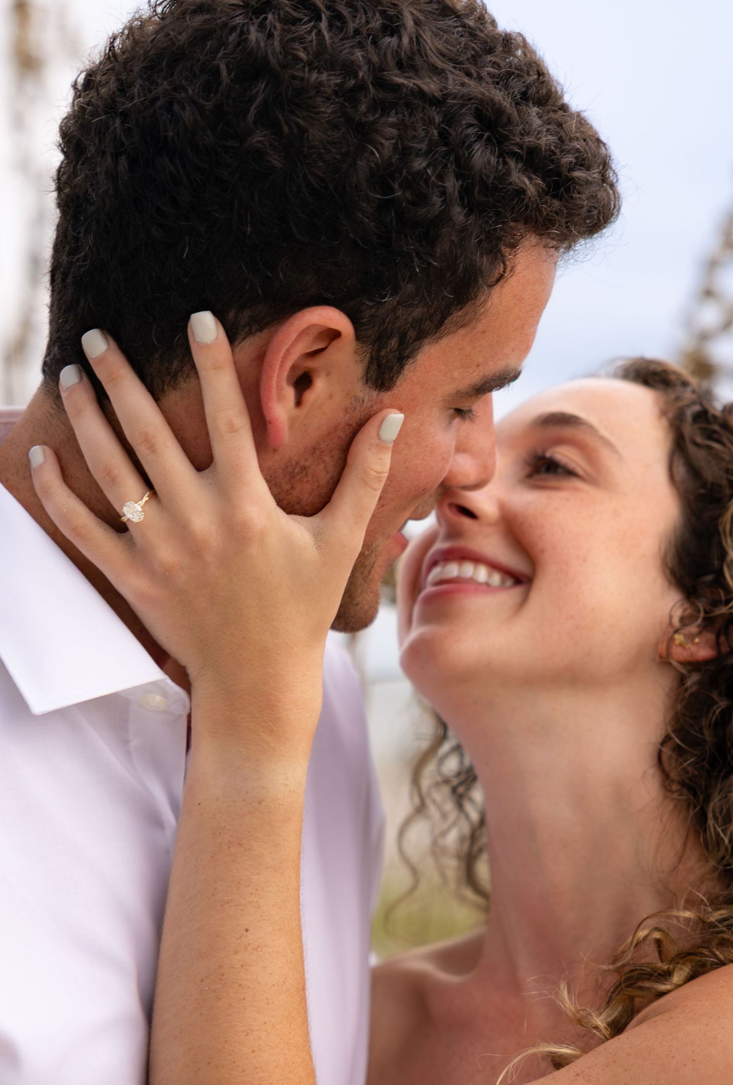 A smiling bride with curly hair and engagement ring tenderly holds her groom's face as they share a romantic moment.