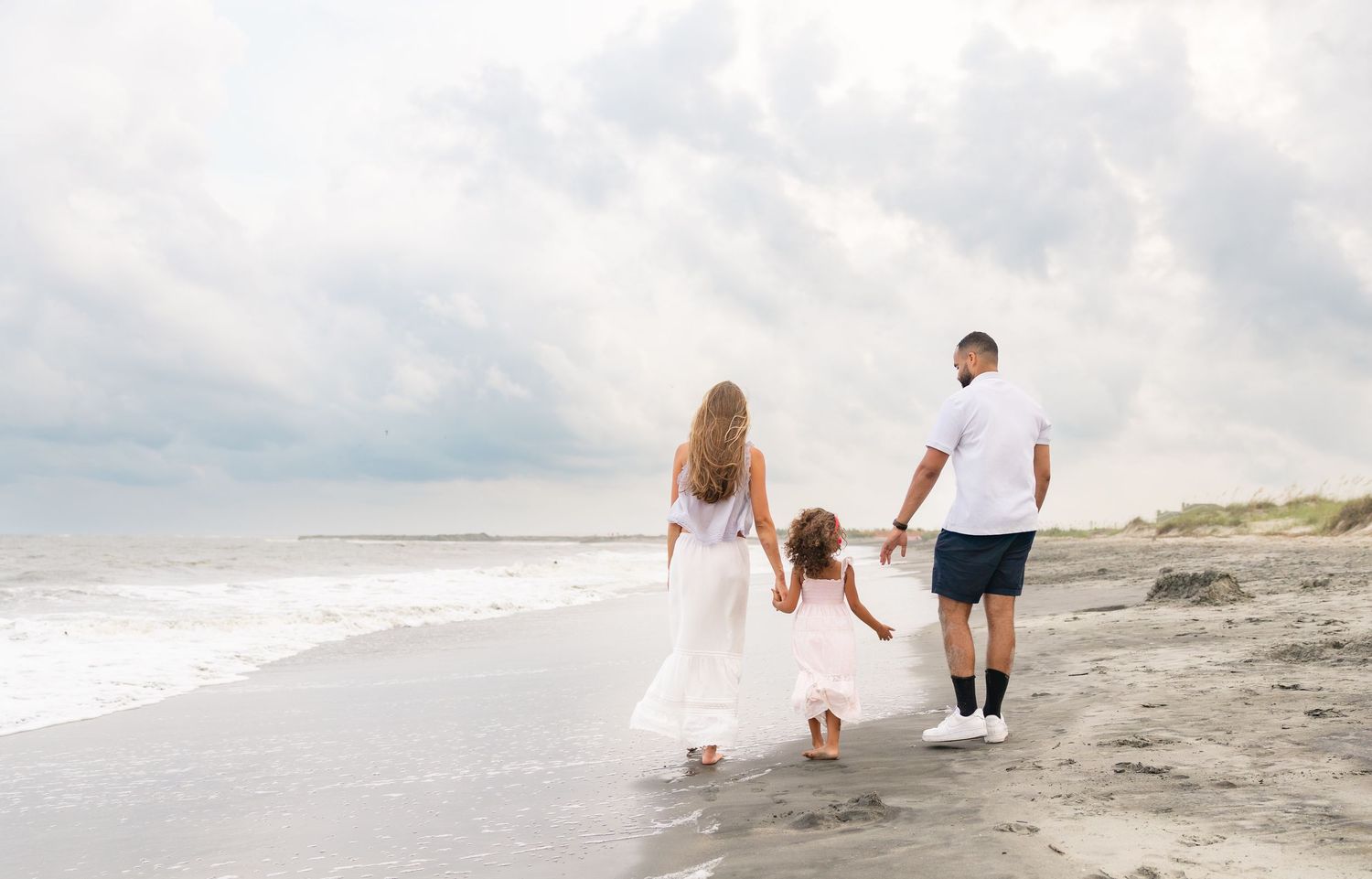 Family of three walking hand-in-hand on a sandy beach, wearing white outfits, with ocean waves and cloudy sky.