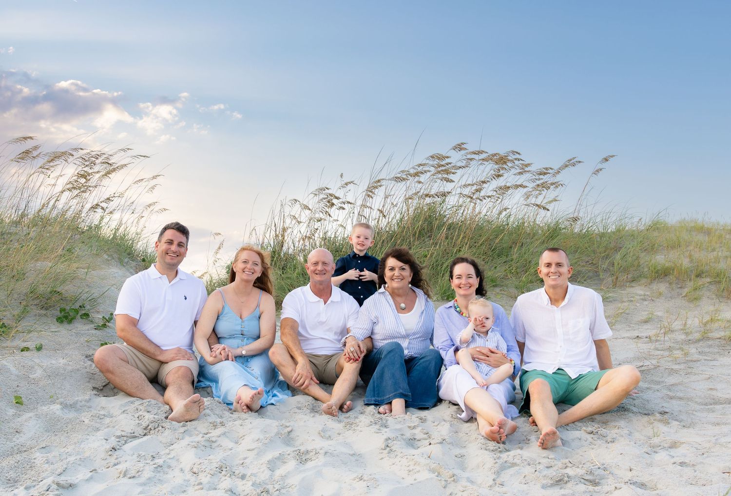 Large family group portrait sitting together on sandy beach dunes with sea grass and blue sky backdrop.