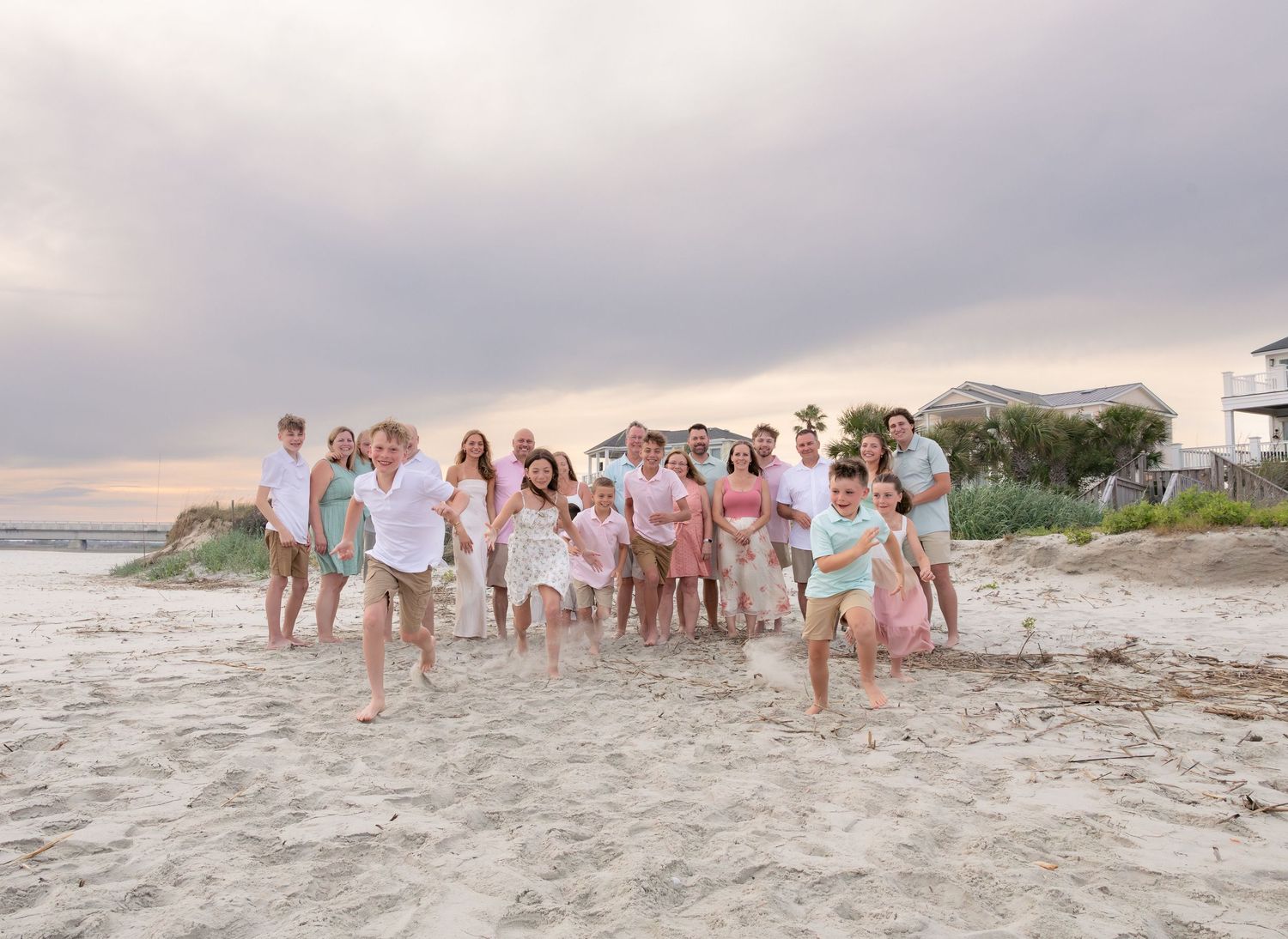 Large family group posing joyfully on a sandy beach at sunset, wearing coordinated pastel outfits near coastal homes.