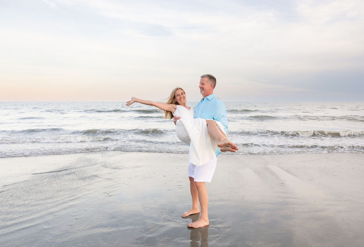 A man lifts a woman in his arms on a sandy beach at sunset, both wearing white and light blue outfits.
