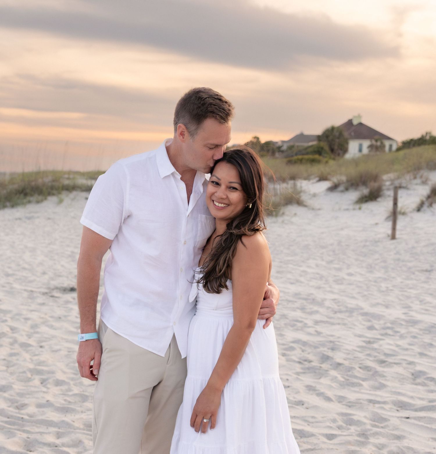 Couple in white outfits smiling on a sandy beach at sunset with dunes and a house in the background.