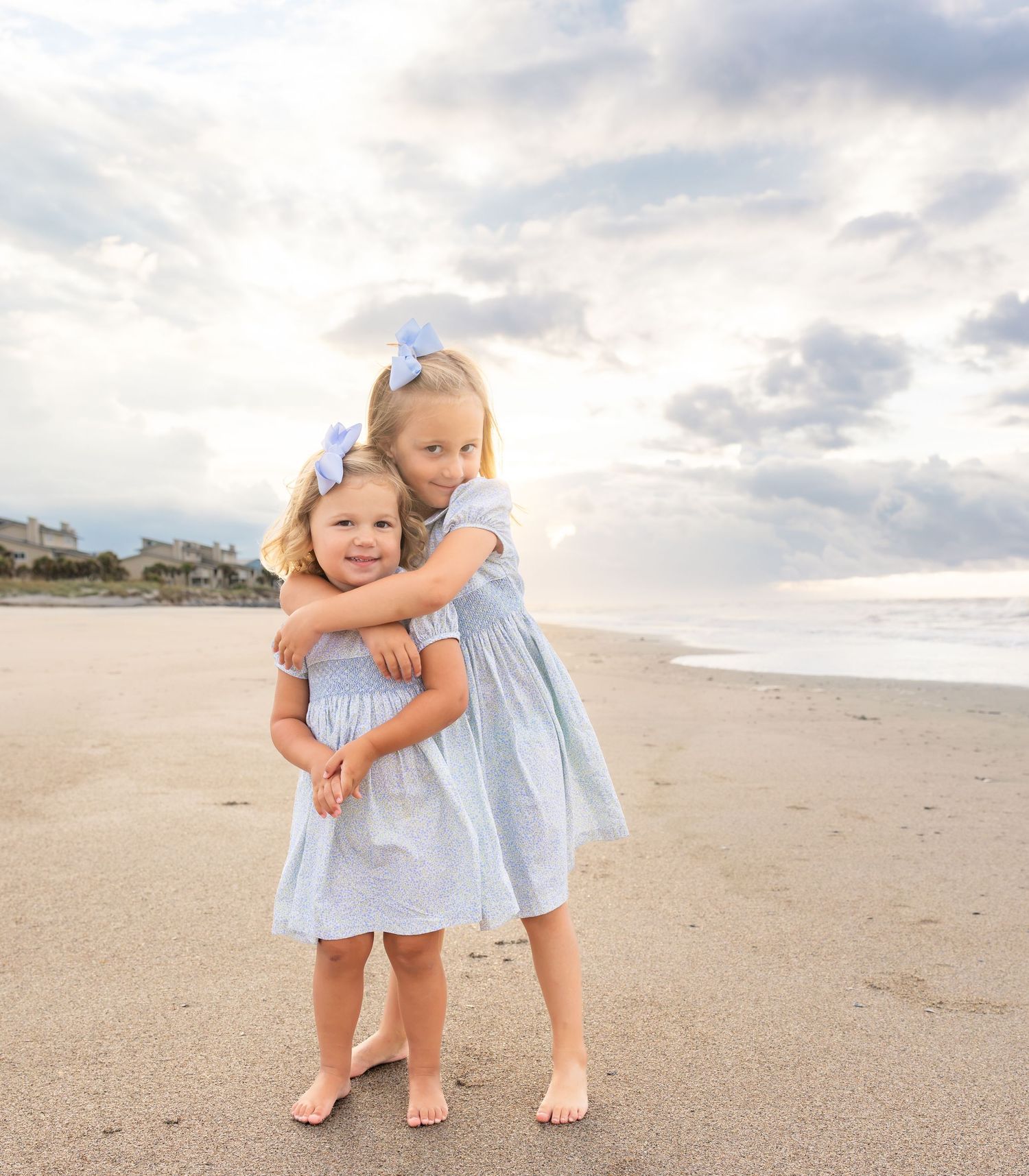 Two young sisters in matching blue dresses hug on a sandy beach with ocean waves and cloudy sky behind them.