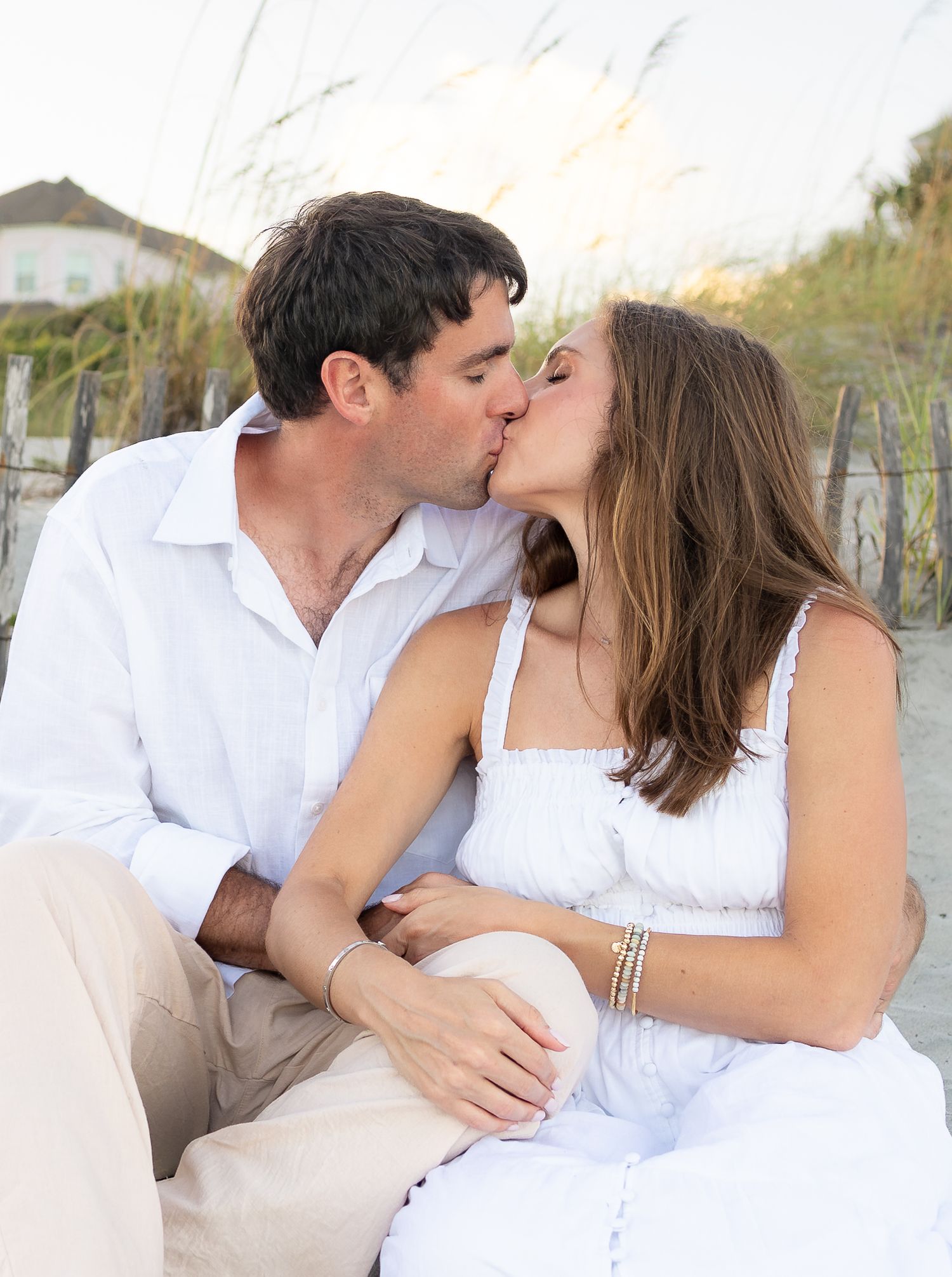 Couple in white outfits sharing a romantic kiss on a sandy beach with sea oats in the background.