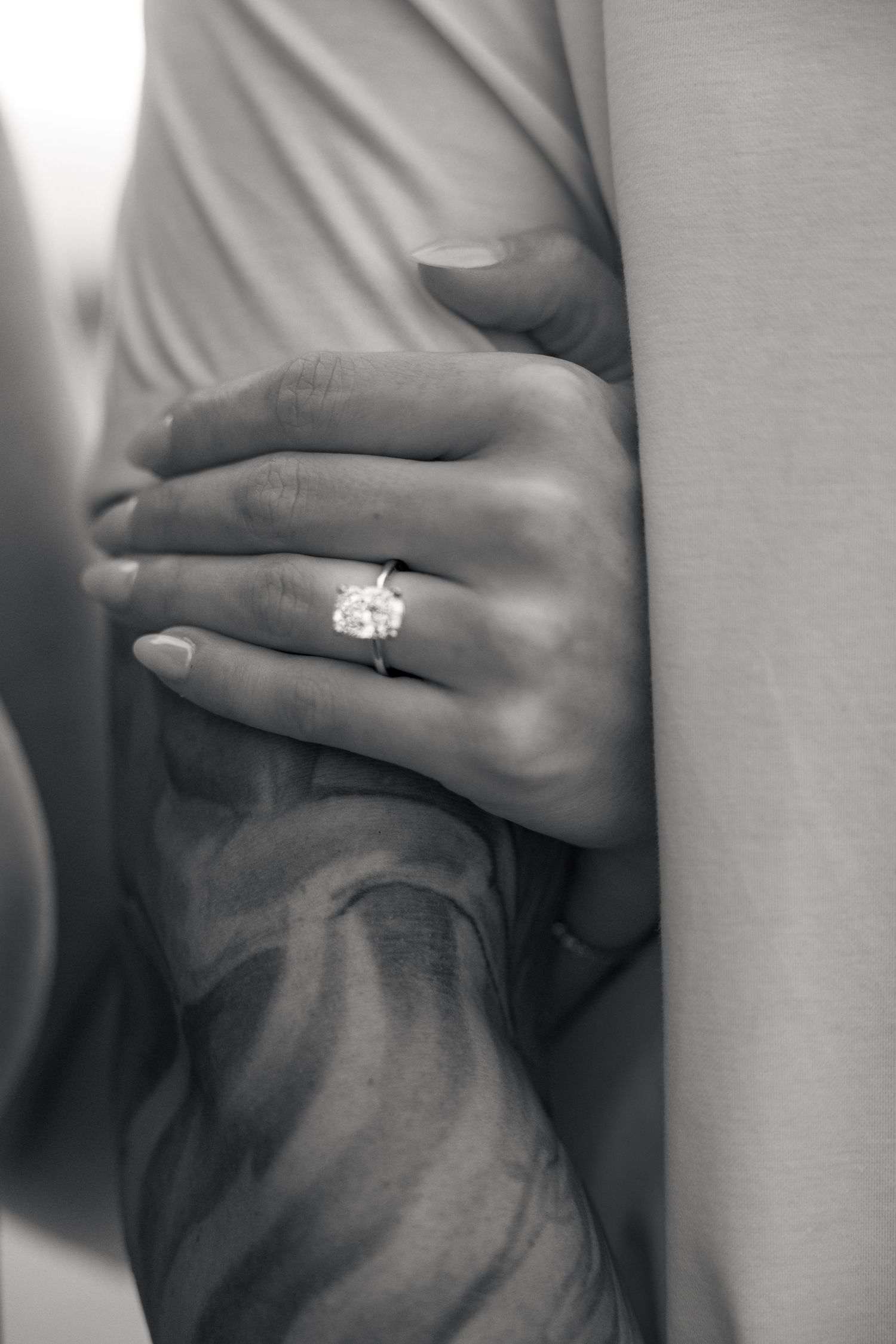 Black and white photo of a woman's hand with a large diamond engagement ring resting on a man's arm.