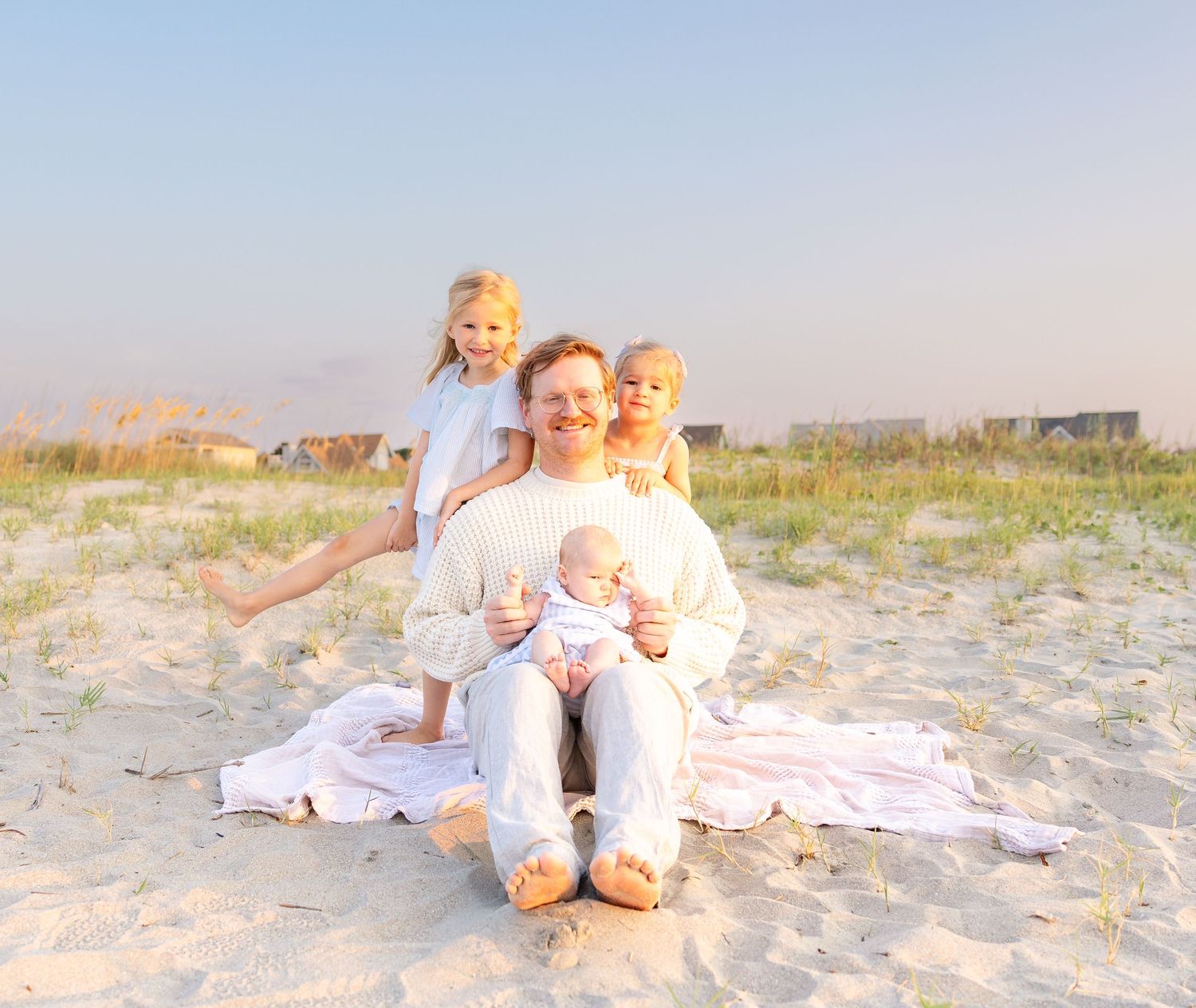 Parent sitting on beach sand with three children, including a baby, during golden hour at a serene coastal setting.