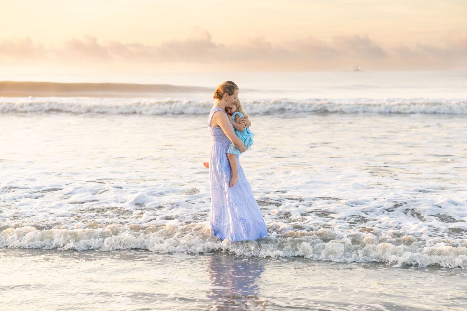 Woman in white dress holding child at ocean shore during golden sunset.