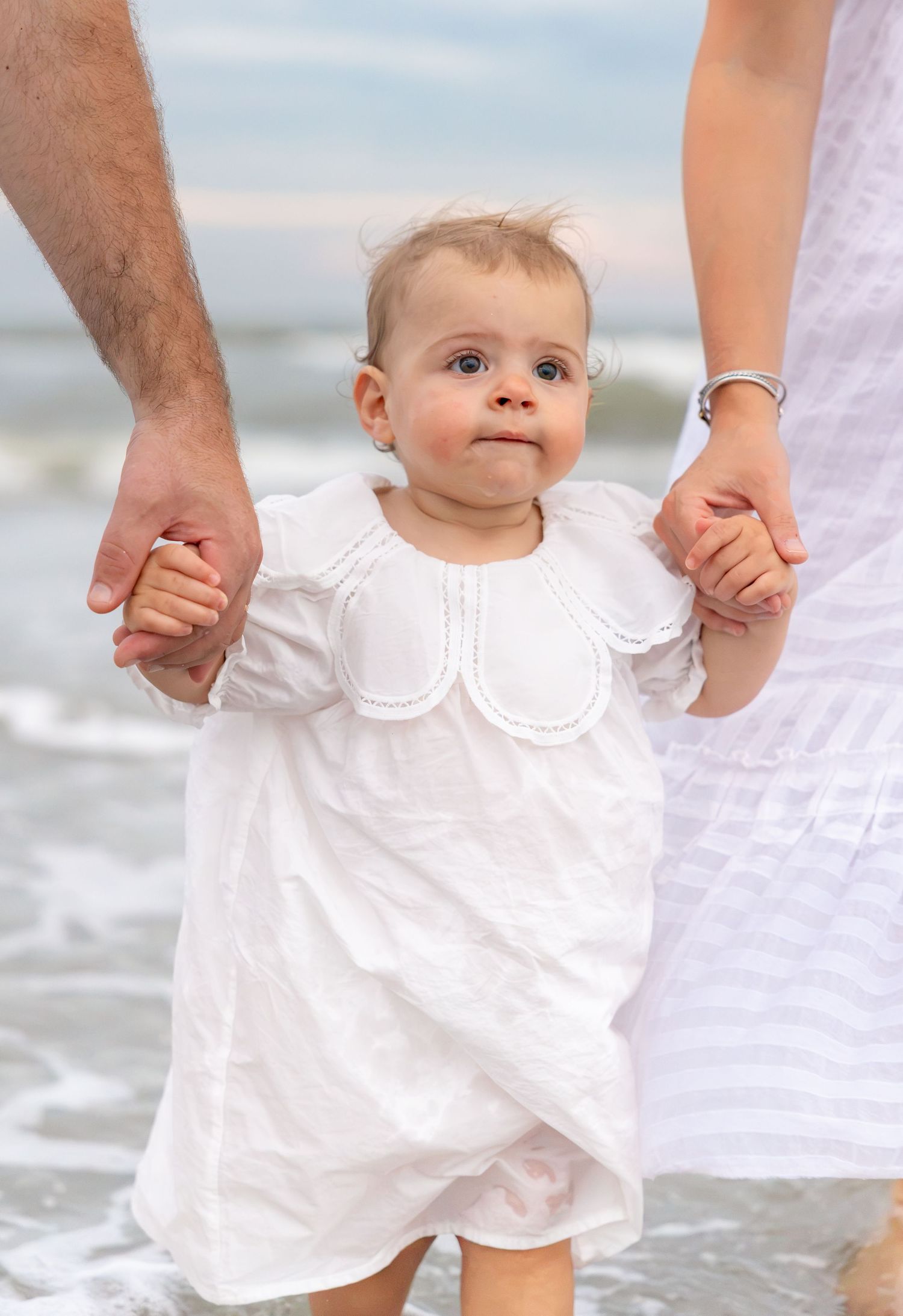 Adorable baby in white dress taking first steps on beach held by parents hands.